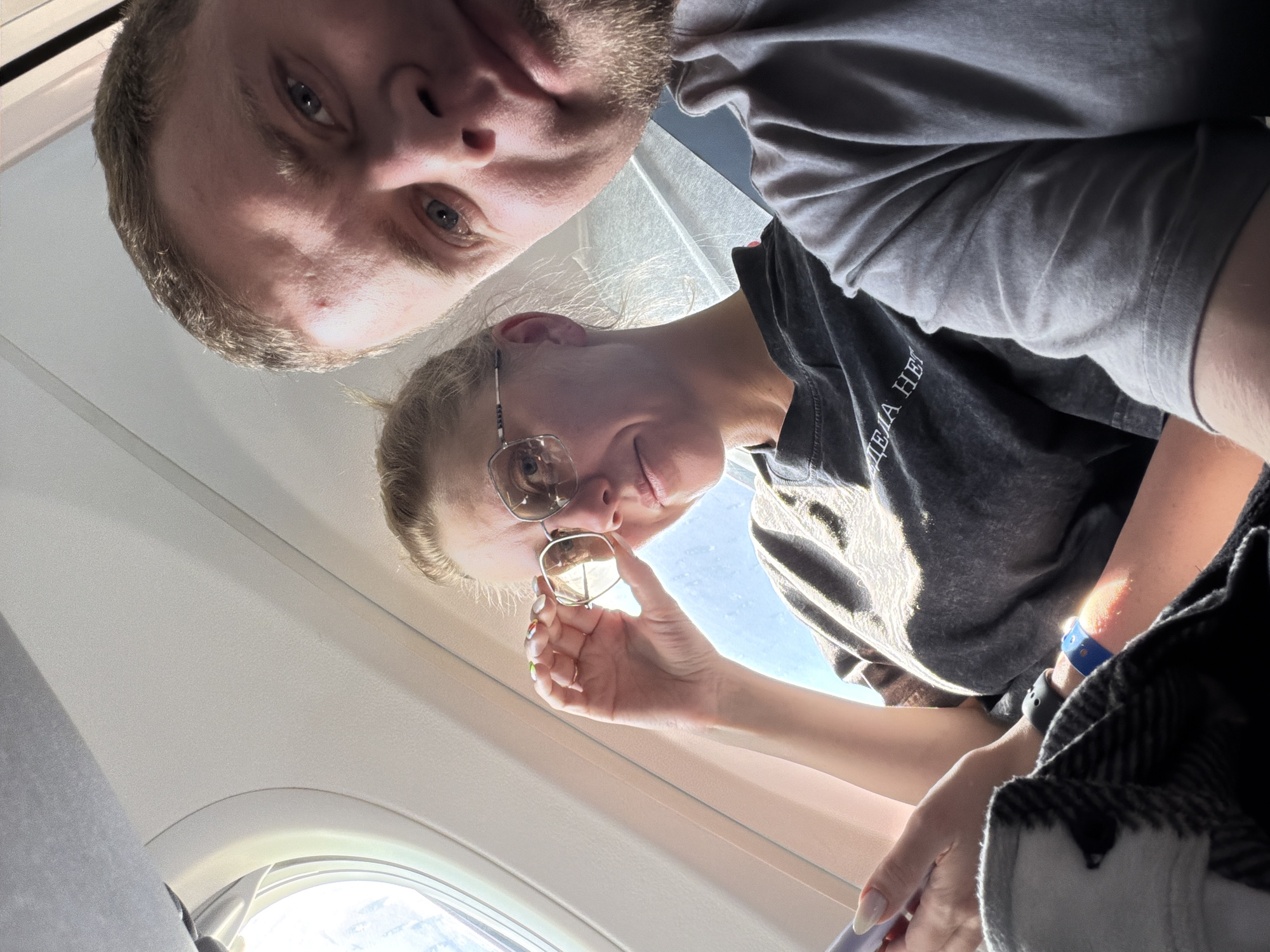 Two individuals are visible in the image. A male with short brown hair and light eyes is situated in the upper left corner. A female with blonde hair pulled back and wearing glasses and a dark t-shirt is in the lower right corner. The female is holding a pair of sunglasses. They appear to be inside an airplane, as indicated by the presence of an airplane window and interior paneling. No specific information to locate the image in Perm, Russia is available.