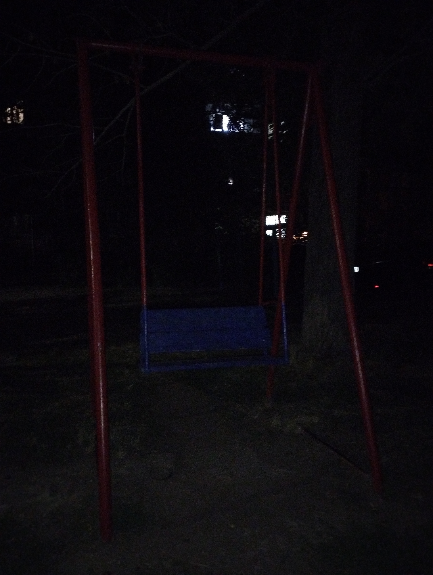 The image shows a swing set at night in an outdoor urban environment. The swing set has a red metal frame with a blue seat. It is located in the city of Belaya Kalitva, Russia. The scene is dark, with some distant lights visible, suggesting it is in a park or residential area. There are no people or animals visible in the image, and no actions or interactions are depicted. The ground appears to be dirt or gravel. The overall atmosphere is still and quiet due to the nighttime setting.