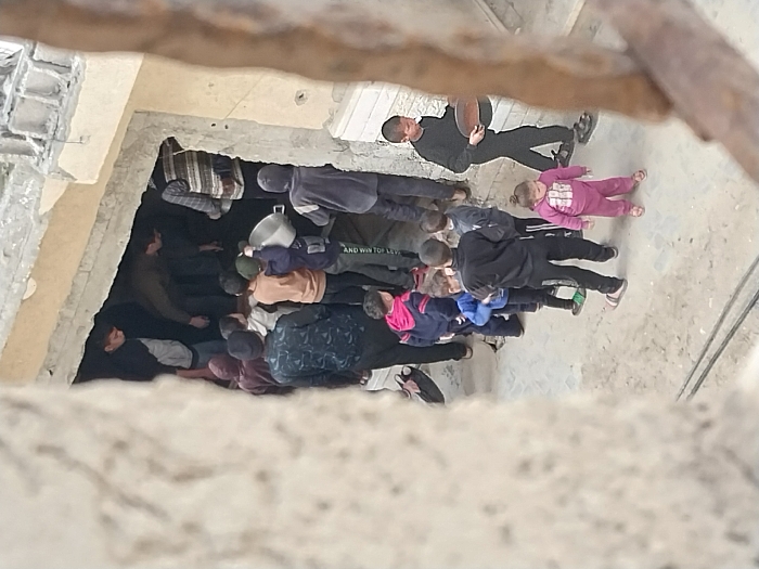 An overhead view in Khan Yunis, Palestinian Territory, depicts a group of approximately 15 individuals, predominantly children and young males, gathered at the entrance of a damaged building. The entrance is a large, irregularly shaped opening in a concrete-colored wall, leading into a dark interior. Visible structural damage includes exposed rebar and rough edges along the top and sides of the opening, and chipped plaster on the building exterior.
A queue of children, aged approximately 4-12 years, forms outside the entrance. One child, wearing a grey hooded top, is positioned towards the center of the line, holding a large, reflective silver pot or bowl. Behind him, other children are visible. To the right of the queue, a male child in a black tracksuit walks away from the opening, carrying a large, round, dark metal tray or pan in his left hand. At the front of the queue, near the opening, several adults or older teenagers are partially visible within the shadowed interior, some bending over. A young female child in a pink tracksuit stands alone to the right, facing towards the left.
The ground surface is a rough, light-colored concrete or packed dirt, with visible dark cables running across it in the foreground. The lighting suggests an outdoor daytime scene.