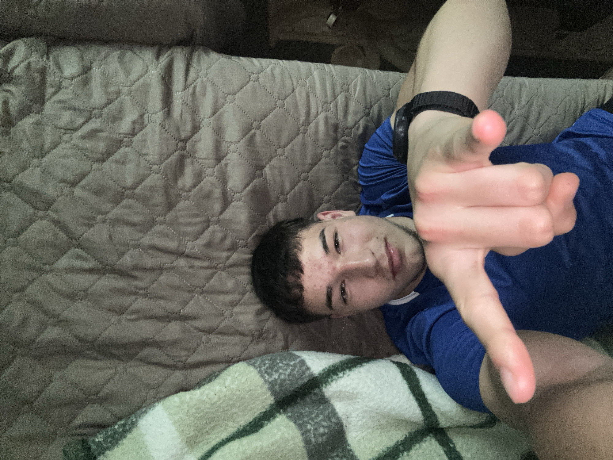 A young man with dark hair is lying on a brown quilted bedspread, partially covered by a green and white plaid blanket. He is wearing a blue shirt and a black watch on his left wrist. He is making a hand gesture with his right hand, holding up his index finger and thumb at a ninety degree angle. The location is unknown and can not be determined from the image.