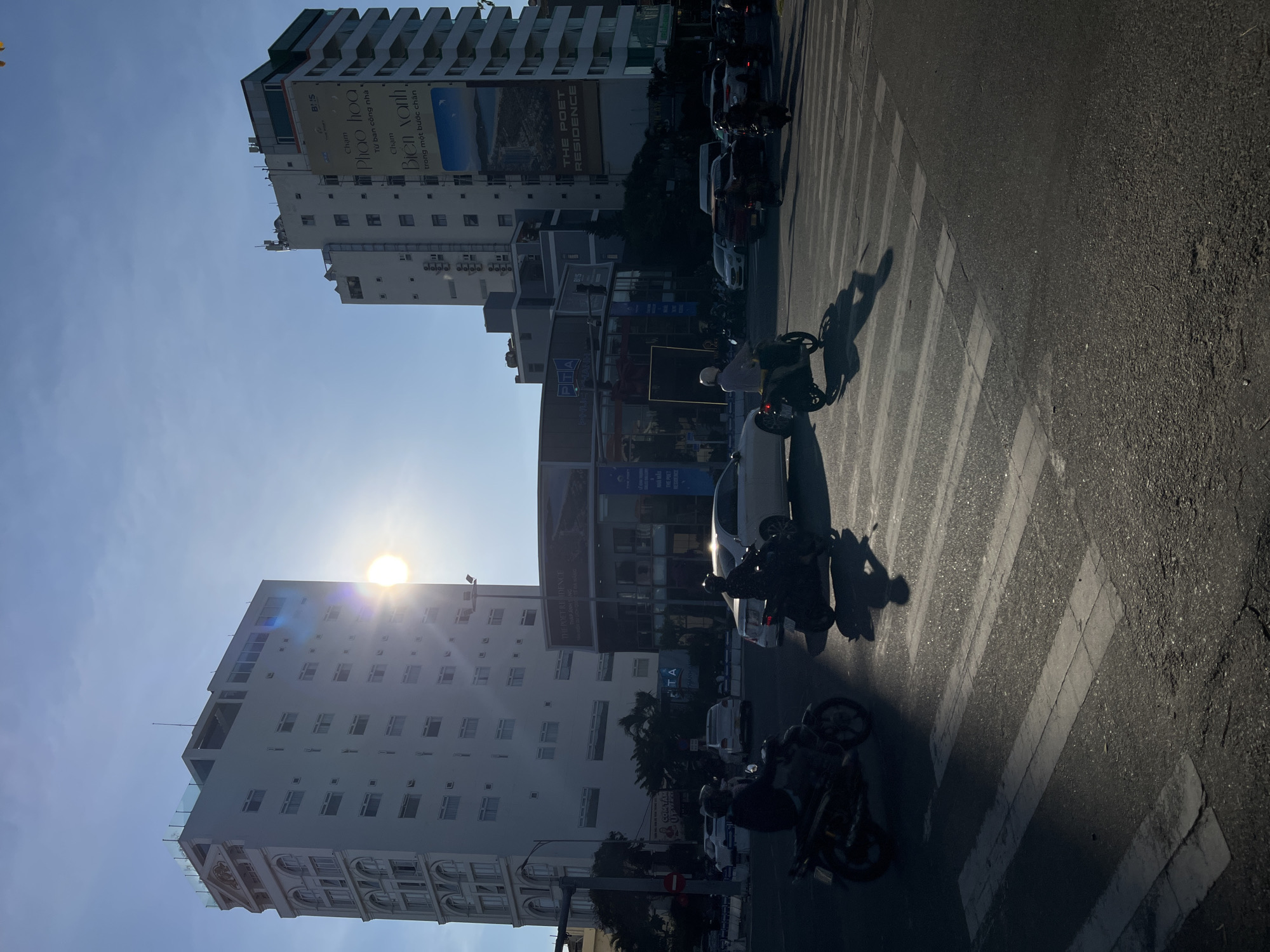 Captured in Đà Nẵng, Vietnam, the image presents an urban street scene with the sun positioned high, causing lens flare. The foreground features a crosswalk with several parked motorbikes casting long shadows, suggesting a sunny morning or afternoon. Buildings line both sides of the street, including a tall white building on the left and a taller building on the right with a large advertisement reading "THE POET RESIDENCE." A building in the center is partly obscured by the sunlight, but signage is visible. Cars are also seen amidst the motorbikes, indicating a busy traffic environment. The overall perspective is from street level.