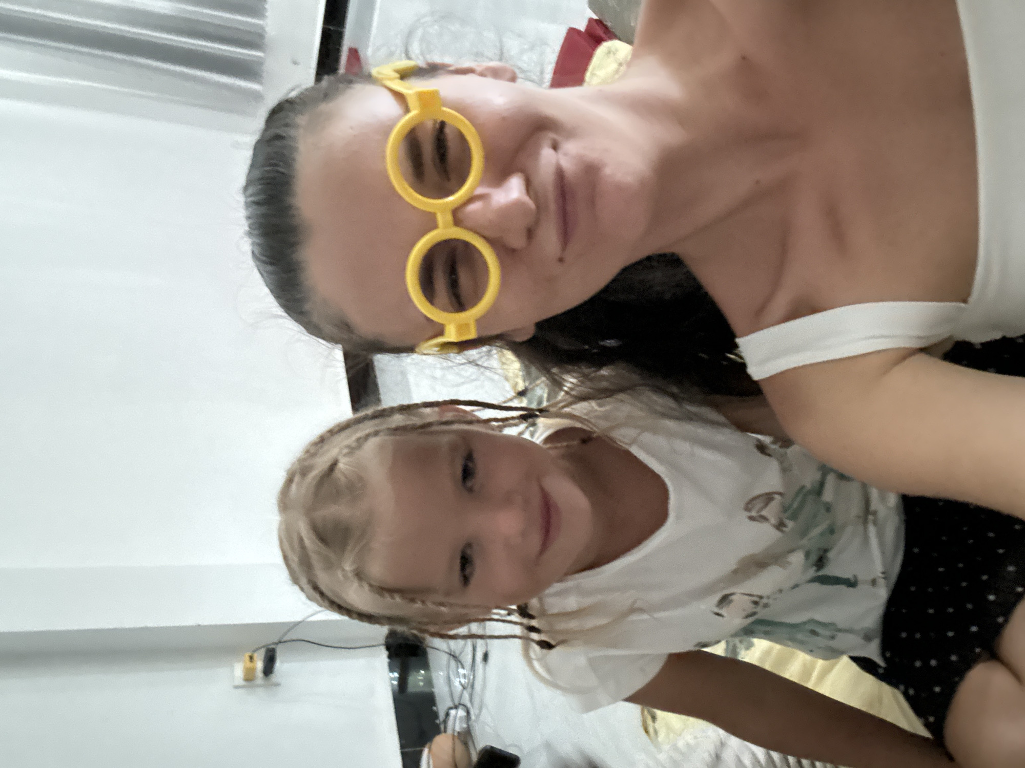 A selfie-style shot captures an adult and a child, both with fair complexions and slight smiles. The adult, wearing whimsical yellow round glasses, is positioned at the top of the frame, partially obscuring the background with their head and upper body. They are wearing a white top. The child, positioned below the adult, also wears a white top with a floral pattern and dark shorts with a polka dot pattern. The child has light hair styled with braids. A white wall with visible moldings is discernible in the upper left corner of the frame, suggesting an indoor environment. A light switch and what appears to be a phone charger can be seen on the wall. The location is Mũi Né, Vietnam.
