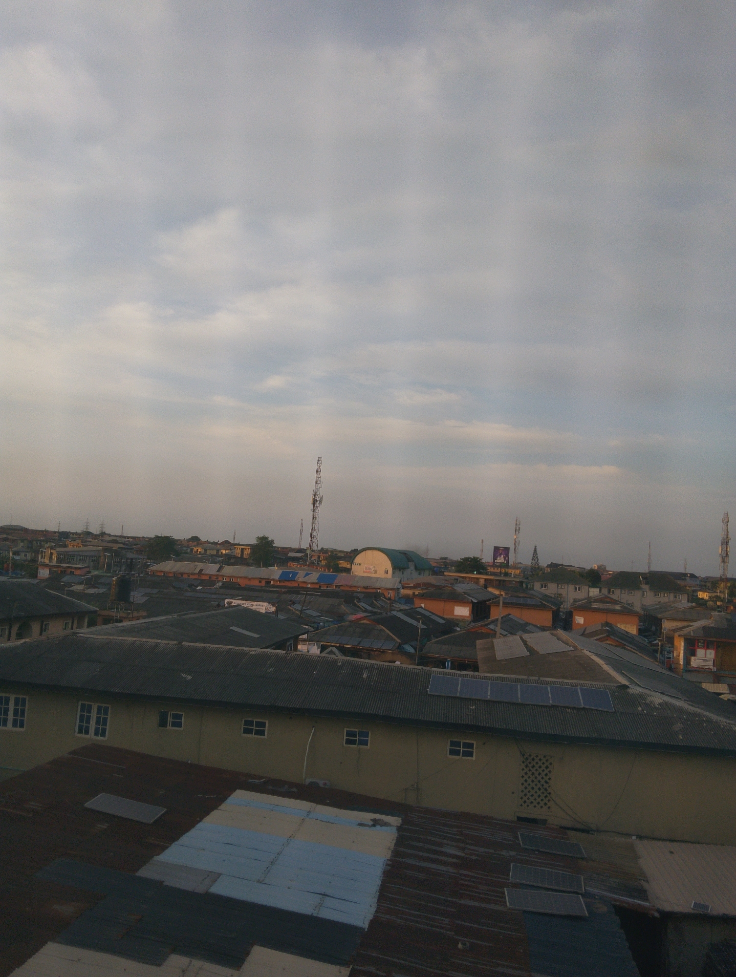 This is an aerial view of a densely populated urban area, likely within Lagos, Nigeria, characterized by a multitude of buildings with corrugated metal roofs. The scene depicts a collection of residential and commercial structures, many of which appear to be closely packed together. Several prominent telecommunication towers are visible in the distance, indicating an active communication infrastructure. The sky is overcast with scattered clouds, suggesting a hazy or potentially rainy atmosphere. Solar panels are visible on some of the rooftops, suggesting an effort towards sustainable energy in this environment. The overall impression is one of a vibrant, built-up urban landscape.