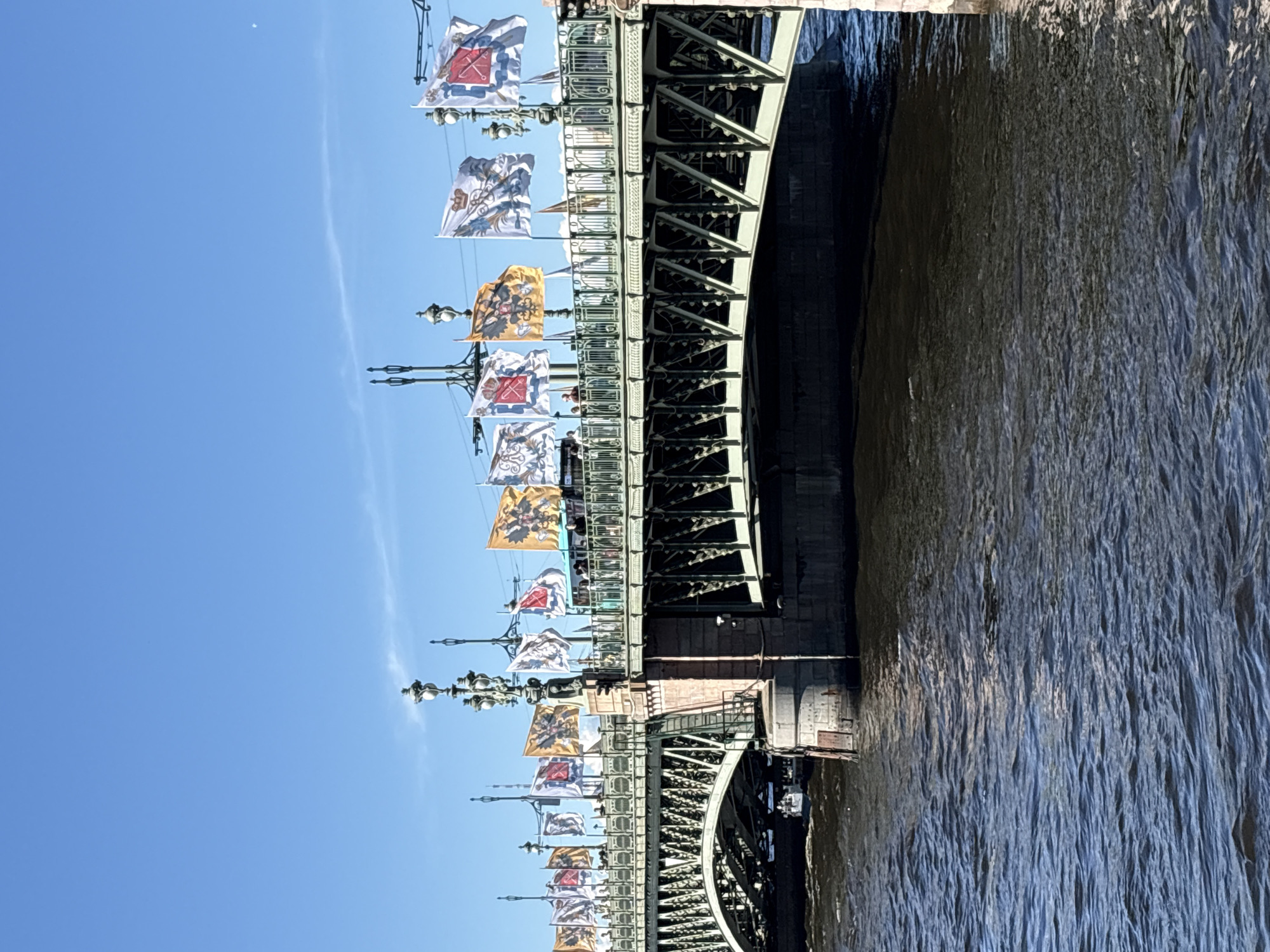 This image depicts a section of the Trinity Bridge (Troitsky Most) over the Neva River in Saint Petersburg, Russia, under a clear blue sky. The bridge features a dark green metal truss structure with prominent arched spans, supported by robust stone piers. Ornate black lampposts with multiple decorative elements and light fixtures are mounted along the bridge's upper deck.
Numerous rectangular flags are displayed from poles attached to these lampposts, appearing to alternate in design. Visible flag designs include a black double-headed eagle on a gold background (the Russian Imperial Standard), a white flag with a crowned emblem and Cyrillic script, and a white flag featuring a red shield or cross-like emblem. These flags are partially unfurled and rippling due to a breeze.
Several pedestrians are visible on the bridge's walkway. A vehicle, partially visible behind the flags, shows a light blue upper section, suggesting a bus or trolleybus traversing the bridge. The river below is a dark blue-grey, exhibiting numerous small ripples and waves reflecting direct sunlight. A small white boat is partially visible passing under one of the bridge's arches on the left. The sky is a bright, unclouded blue, with a faint, wispy cloud streak discernible in the distance.