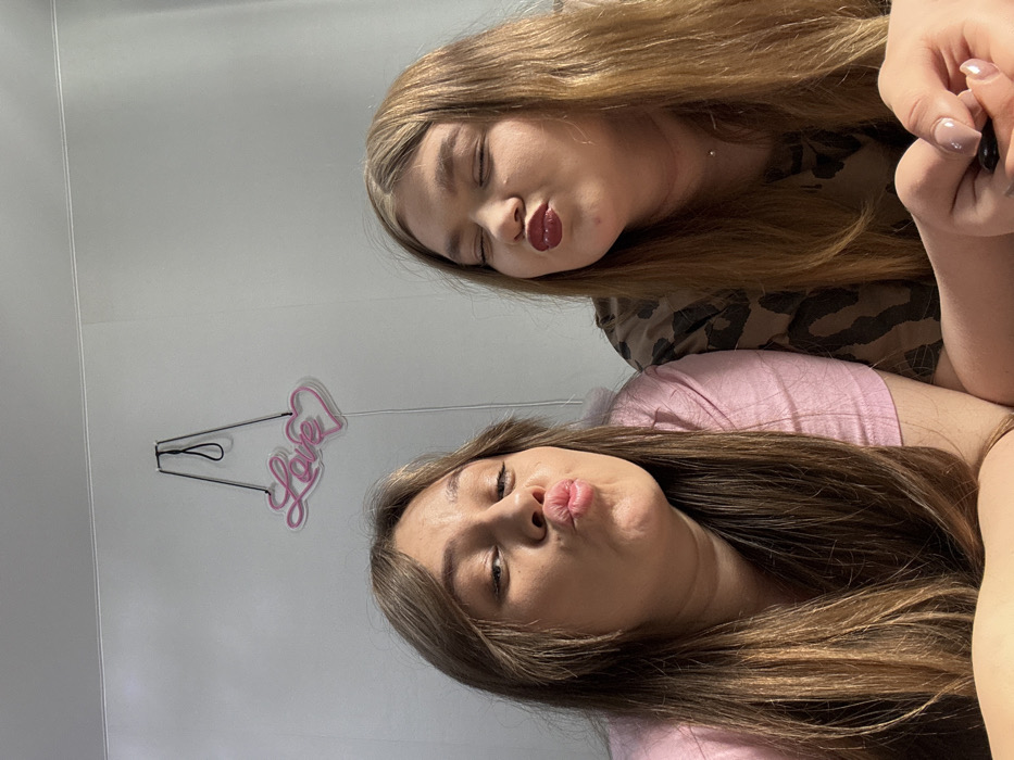 Two young adult females are depicted posing for a photograph in an indoor setting in Chaikovsky, Russia. The individual on the left, with long light brown hair and a pink short-sleeved top, faces forward with lips pursed. The individual on the right, with long reddish-brown hair and a camouflage-patterned short-sleeved top, has closed eyes and also presents a pursed-lip expression, marked by dark red lipstick. A minimalist silver necklace is visible on the right subject. Her right hand, featuring light pink manicured nails, is visible in the lower right foreground, holding a dark object. A pink neon sign, displaying the word "Love" alongside two hearts, is mounted on the plain light grey wall behind the subjects. The scene is evenly illuminated.