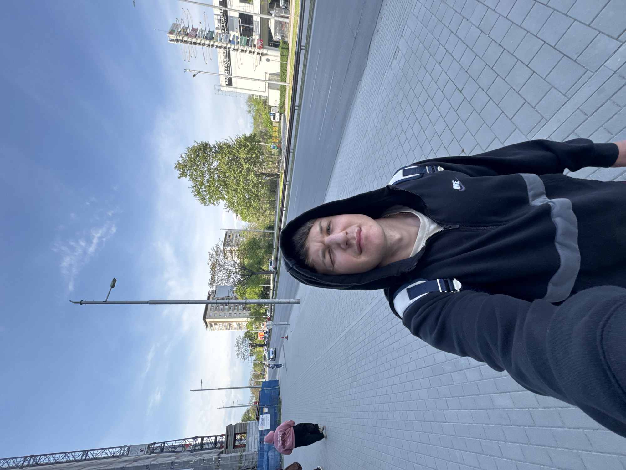 A selfie captured in an urban setting during the daytime. The primary subject is a young person, likely male, in the foreground, wearing a black hooded zipper jacket with a visible "NIKE" logo on the left chest and grey stripe details. A cream-colored shirt is visible underneath the jacket. He has a neutral to slightly playful expression and looks directly at the camera. A strap from a bag or backpack is visible on his right shoulder.
The person stands on a grey tiled sidewalk next to a multi-lane paved road. In the background, various buildings line the street, including one large white building with multiple colorful banners and signs displaying partially visible text such as "MARTINEZ" and "ZENIT". Another building, possibly older residential, is visible further down the street, and a construction site with scaffolding and blue temporary fencing is present in the lower left, where some unreadable text is also visible.
Green trees are scattered throughout the scene, and several streetlights stand along the road. The sky is clear blue with some wispy white clouds, indicating sunny weather. A person in a pink hoodie and dark pants walks away from the camera on the sidewalk in the mid-ground. The overall scene depicts a busy urban street environment, likely captured in Plovdiv, Bulgaria.
