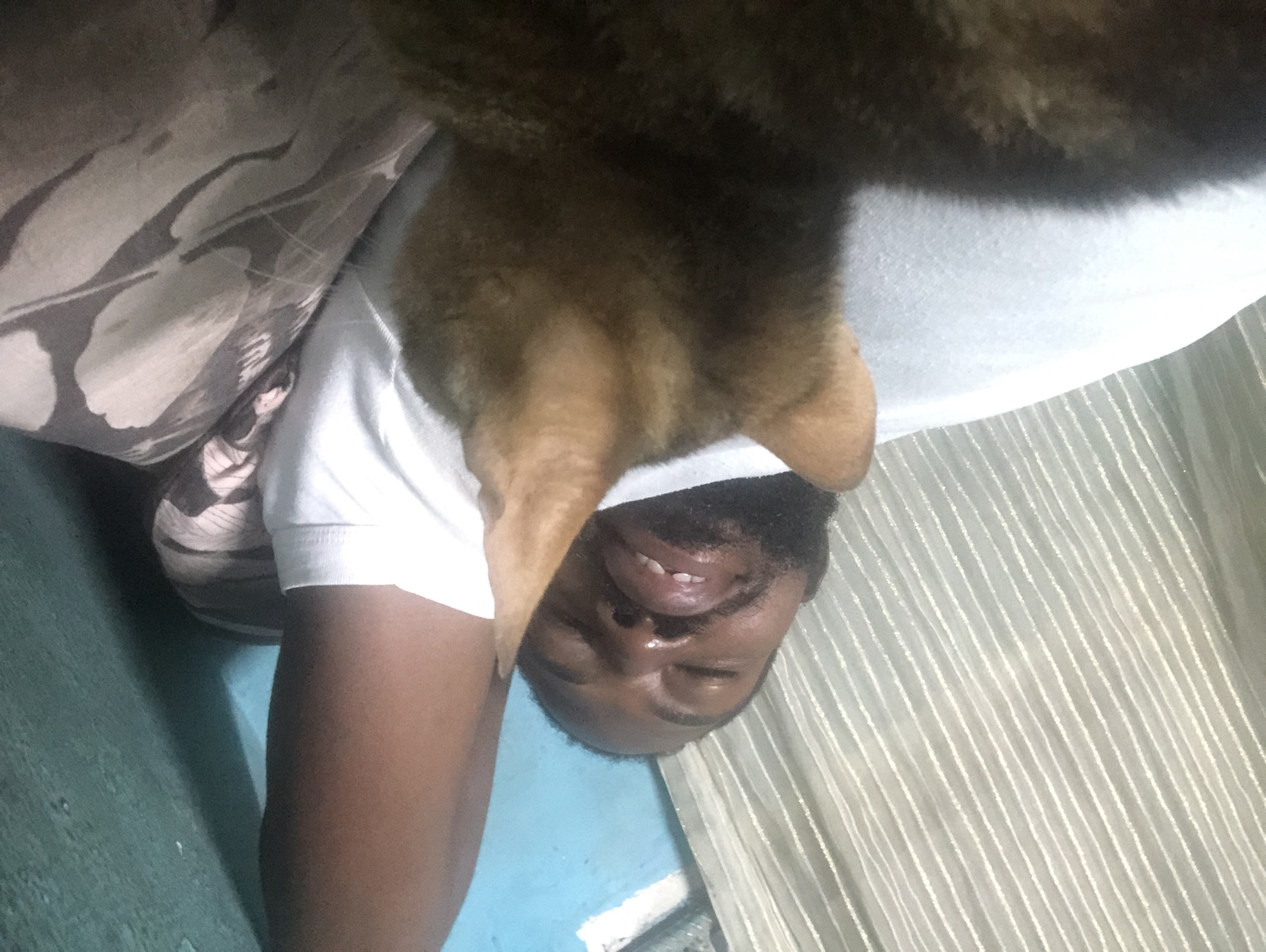 A man is lying down with his eyes closed, his head tilted back. He has a beard and is wearing a white t-shirt. A cat is in the foreground, facing away from the camera and towards the man's face, with its back and head visible. The background consists of a striped curtain on the left and a teal wall on the right. The overall impression is of a relaxed, intimate moment between the man and his pet. The scene is likely indoors, within a residential setting in Lagos, Nigeria.