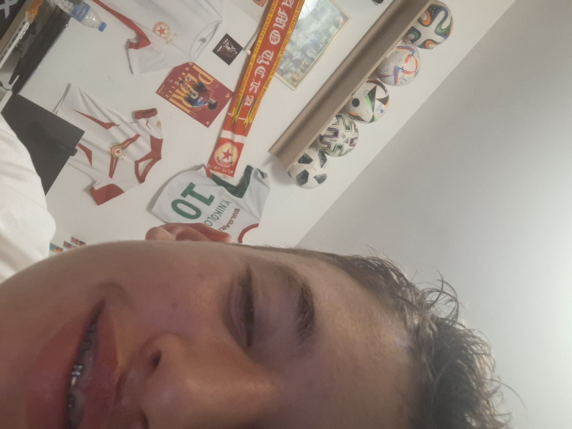 The image displays a close-up, upward-angled selfie of an individual taken in an interior room in Isperih, Bulgaria. The subject's face, from the nose down to the chin on the right side, is visible, showing a partially closed or squinting right eye and a mouth with visible braces. Brown, slightly disheveled hair covers the forehead. The background features a white wall extensively decorated with football-related items. A wooden shelf runs across the upper left, displaying a line of at least five soccer balls, each with distinct patterns. Below the shelf, a framed picture is partially visible, next to a red and white scarf inscribed with "АМО ЦСКА" and featuring a red star emblem. Further down, two white t-shirts with red accents and the same star emblem are displayed, alongside a red poster that reads "PEDRI GONZALEZ". On the right side of the wall, a white football jersey with "BULGARI Y. NIKOLO" and the number "10" in green text is visible, positioned above another white t-shirt with red accents and a star emblem. A plastic water bottle and a portion of a keyboard or desk are visible at the bottom left of the frame.