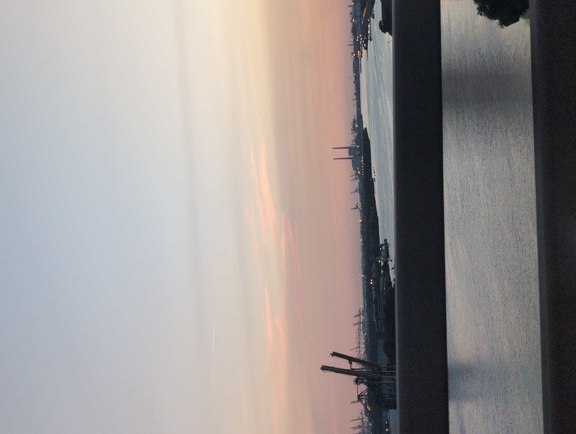 The image shows a wide view of a harbor area in Oudalle, France, during what appears to be either sunrise or sunset. The sky is a gradient of soft colors, transitioning from pale blue at the top to pale pink and orange near the horizon, with faint wisps of clouds.
In the foreground, a dark, blurred horizontal structure, likely a railing or barrier, divides the image. Below this, a body of water, possibly a canal or river, reflects the colors of the sky. The water's surface is calm, with subtle ripples.
The midground reveals a busy industrial port. Prominent features include large cranes with tall, skeletal structures, characteristic of port loading equipment. Several ships or boats are visible on the water. In the distance, industrial buildings and structures, including what appear to be power plant smokestacks, dot the landscape. Lights are visible along the waterfront, suggesting ongoing activity despite the low light.
The overall atmosphere is one of industrial activity set against a tranquil, colorful sky. The precise location is identified as the city of Oudalle, France, known for its port facilities. There are no discernible people or direct human interactions depicted in the image. The focus is on the vast scale of the industrial infrastructure and its natural surroundings.