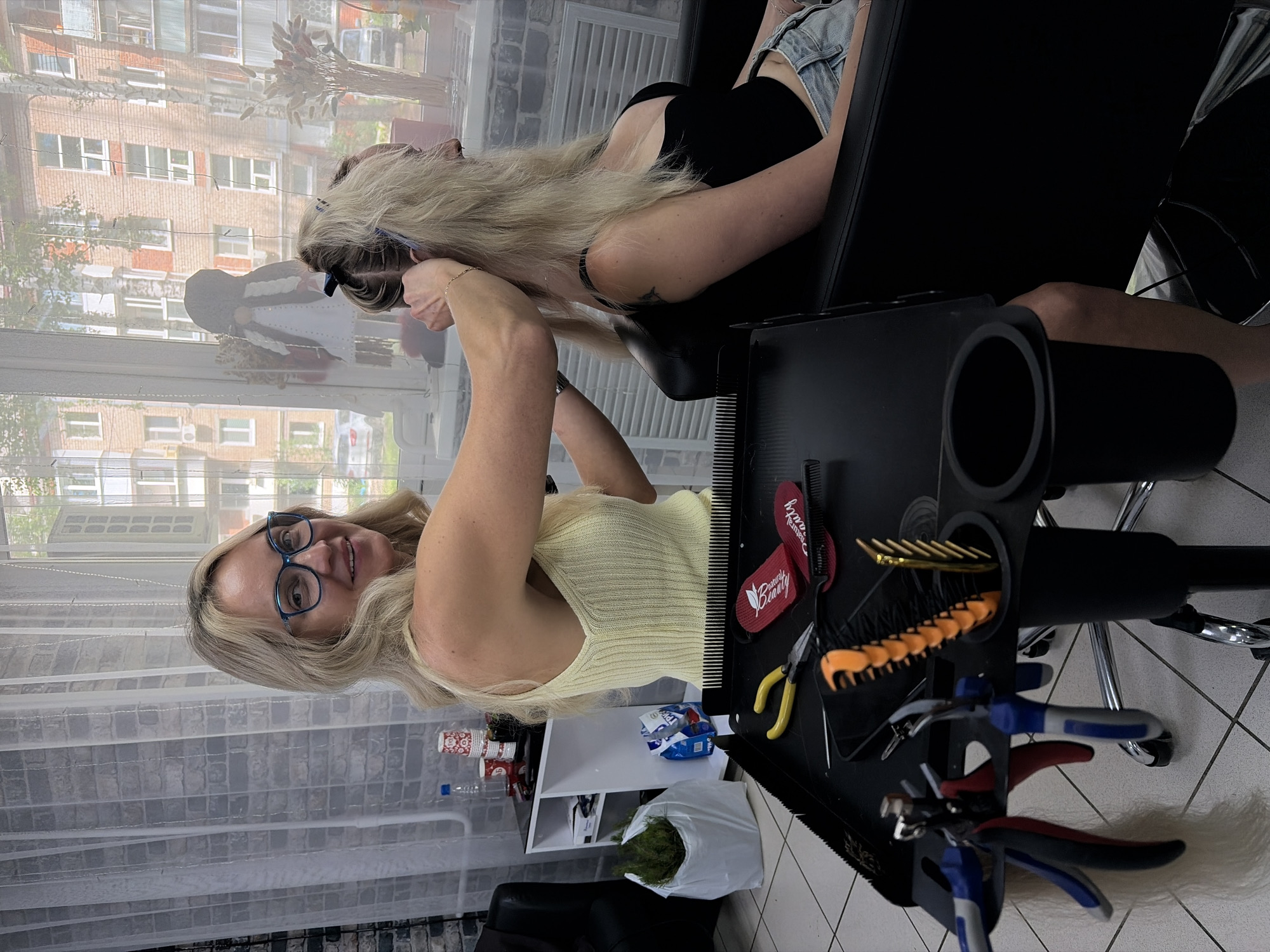 A blonde woman with glasses is in a salon, her hair being styled. She is wearing a yellow knitted top and is looking towards the camera with a smile. In the background, a window shows apartment buildings, suggesting an urban setting, possibly in Chaikovsky, Russia. A trolley with hairdressing tools like combs and scissors is positioned in the foreground, and a black leather chair, likely for a client, is visible to the right.