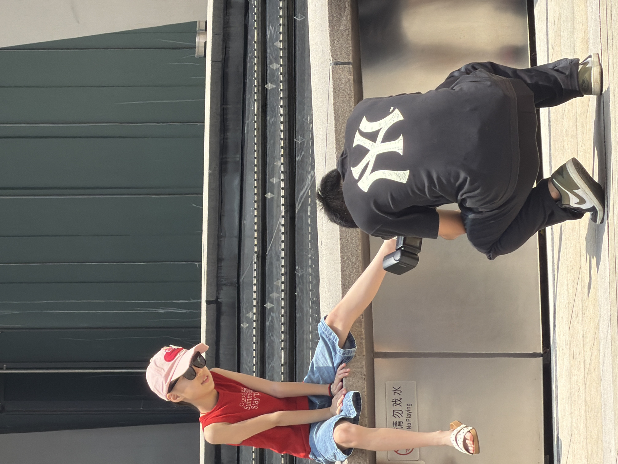 In Yuzhong District, China, a young person wearing a red tank top, denim shorts, a pink baseball cap, and sunglasses is sitting on a concrete ledge. Their left leg is extended forward. A second person, wearing a black t-shirt with a white "NY" logo and dark pants, is crouched down in front of the seated person, holding a camera with a flash attachment. The background shows a dark green metal structure and tiered concrete steps. A sign on the ledge reads "请勿戏水" and "No Playing." The ground is a light-colored paved surface.