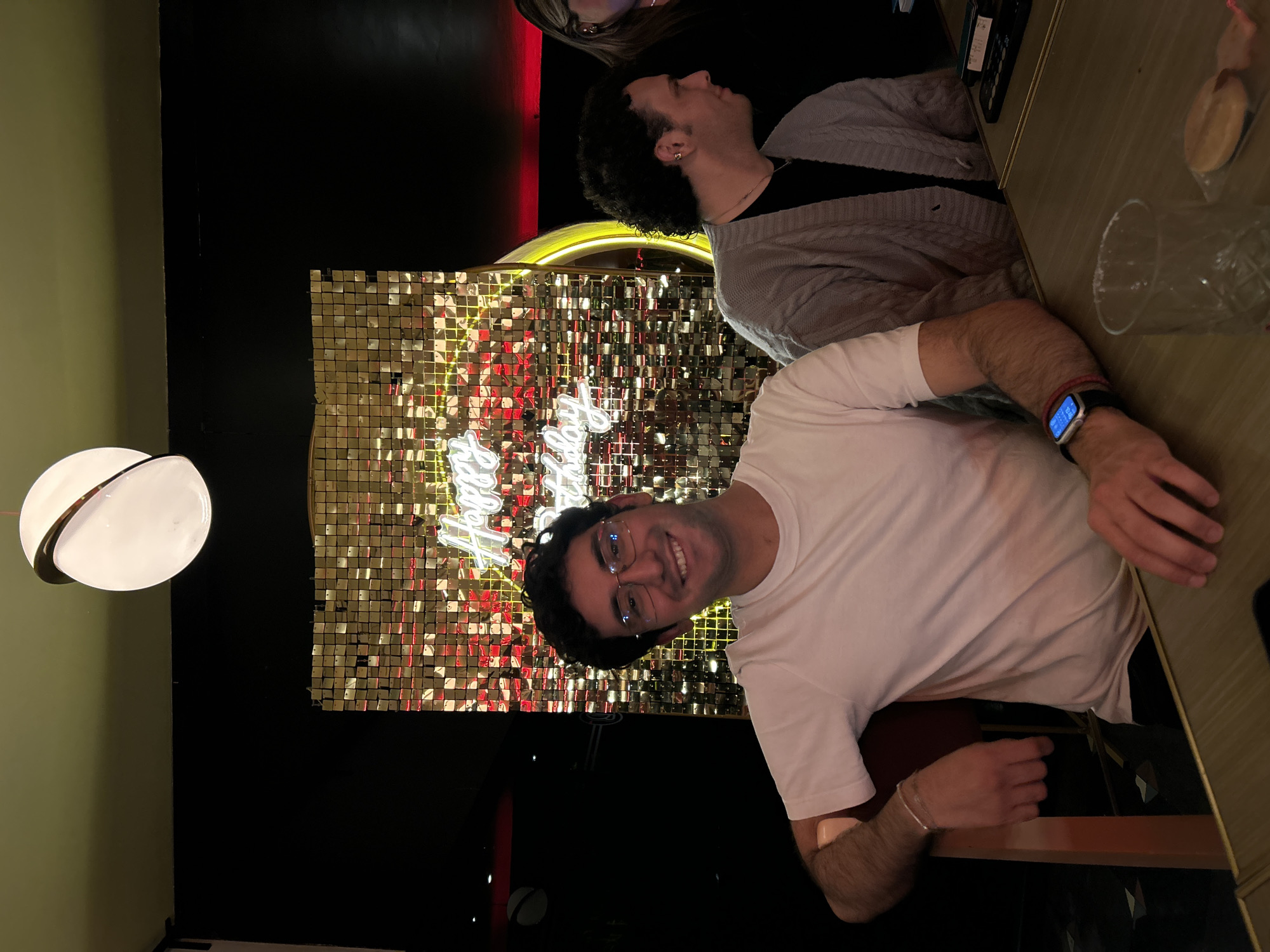 The image depicts two men seated at a table in an indoor setting with a festive atmosphere. The man in the foreground is wearing a white t-shirt, glasses, and a smartwatch, and he is smiling at the camera. The second man is wearing a grey sweater and appears to be looking upwards or towards the side. Behind them, there is a wall decorated with shimmering gold square tiles and a neon sign that reads "Happy Birthday". The setting is dimly lit, with a modern white globe light visible on the left side. There is a glass on the table near the right edge of the image, along with some indistinct objects. Based on the decor and likely celebratory message, it might be a bar or restaurant interior. There is no clear evidence in the image to confirm the location is in Puebla, Mexico.