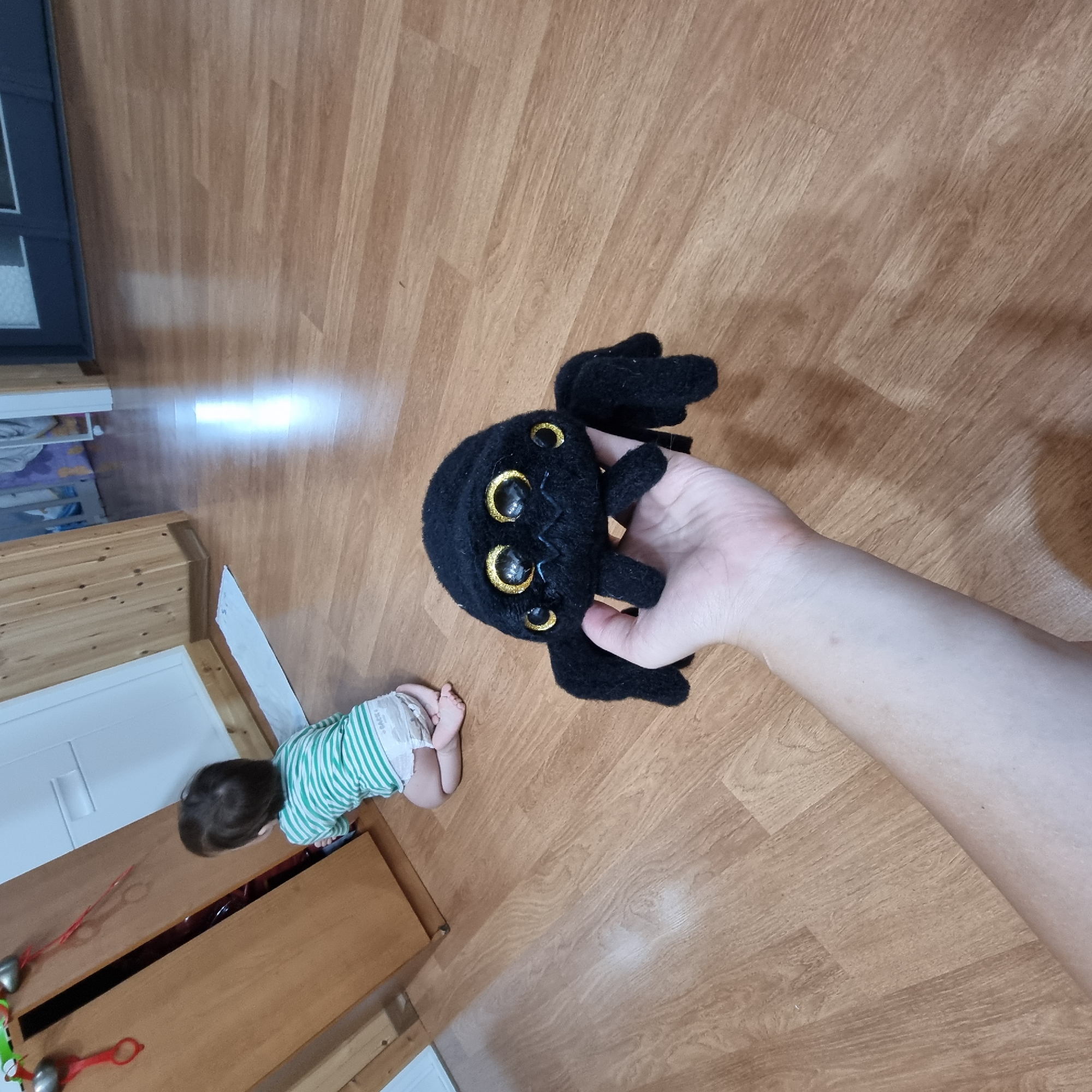 In Dunnae-myeon, South Korea, on a wooden floor, a person's hand holds a black plush spider toy. The spider has large, yellow and black eyes and eight black plush legs. In the background, a baby wearing a green and white striped shirt and a diaper kneels and looks into an open wooden cabinet. The cabinet has red decorations hanging inside.