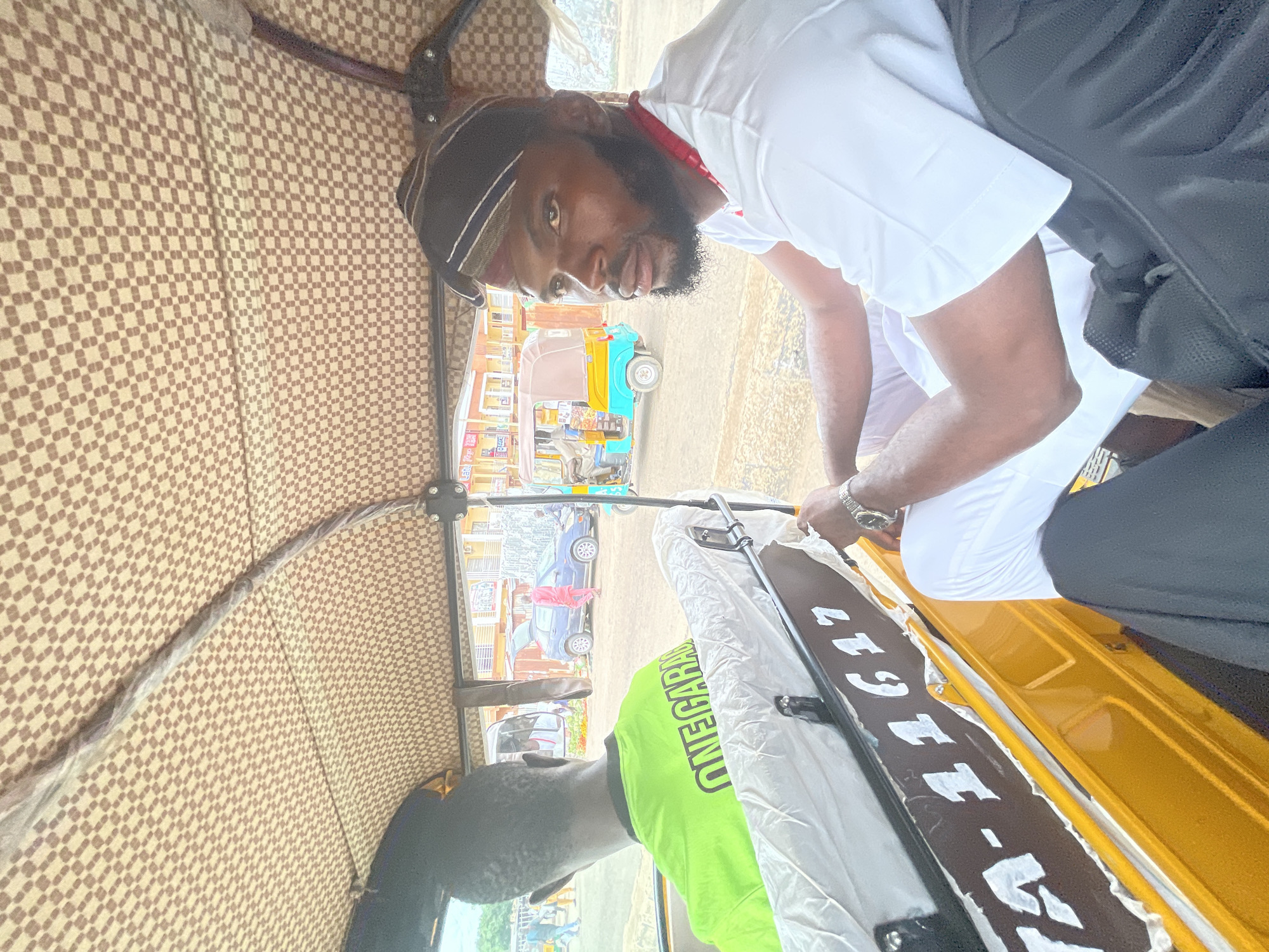 This image presents a detailed view from inside a yellow auto-rickshaw (Keke Napep) on a street in the city of Jos, Nigeria.
In the foreground, on the right, a dark-skinned man with a full beard and mustache is seated, looking towards the left side of the frame with a neutral expression. He wears a white t-shirt, white trousers, and a dark-striped cap with bands of white and brown. A red beaded accessory or lanyard is visible around his neck. A metallic wristwatch is on his right wrist.
To the left, the back of the auto-rickshaw's driver, a dark-skinned man with short dark hair, is visible. He is wearing a neon green t-shirt with the word "ONEGARA" (likely part of "ONEGARAGE") printed in black capital letters across the back.
The auto-rickshaw's ceiling features a brown and beige checkered pattern. The back panel of the passenger compartment, below the driver's seat, is black with white lettering and numbers "TA-11617," partially obscured by white material, possibly a plastic cover or worn upholstery.
The background depicts a bustling street environment. A blue sedan with its trunk open is parked on the left side of the street, with a person in a vibrant pink or red outfit walking near it. Further down the street on the right, another light blue and yellow auto-rickshaw is visible. Several other individuals are present on the street, some walking, others near buildings. Low-rise commercial buildings line the street, with one partially visible sign reading "Let's." The ground appears dusty, and the sky is bright and overcast.