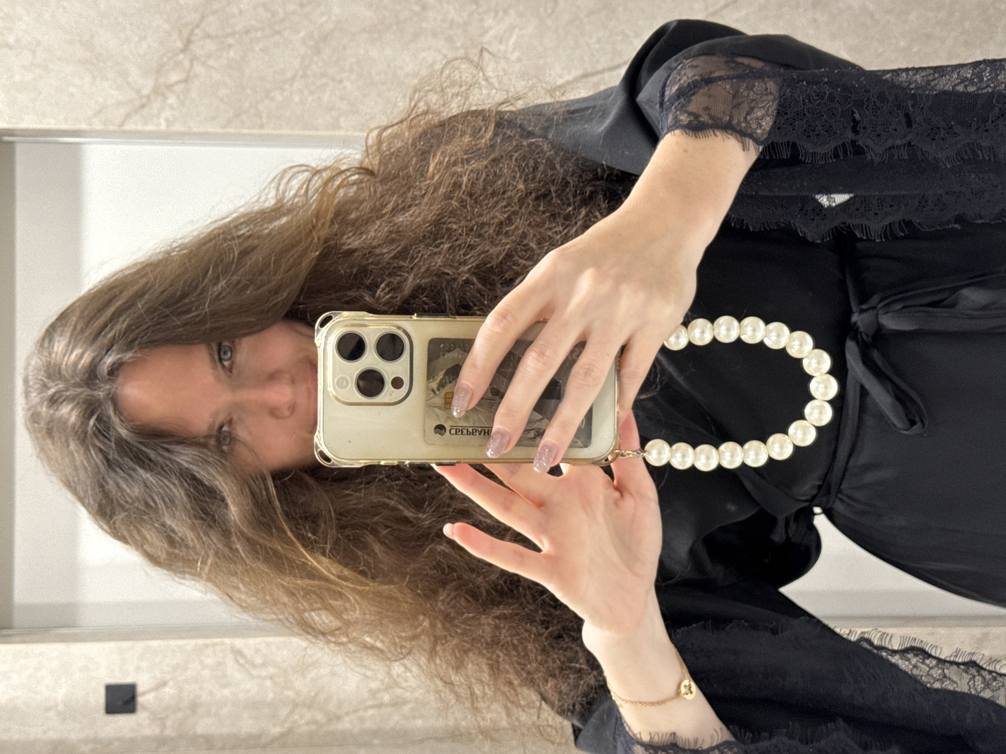 A mirror selfie captures a woman with long, voluminous, auburn-brown wavy hair and blue eyes, reflected sideways. She holds a smartphone, encased in clear plastic, with both hands. Her nails are manicured with glittery polish. A pearl strap, forming a necklace-like loop, is attached to the phone. A small gold bracelet with a circular charm adorns her left wrist. She is wearing a black garment, possibly a robe or top, featuring elegant black lace trim on the sleeves. A credit card is visible behind the phone, held within the clear case, displaying the Cyrillic text "СБЕРБАНК". The background reflection shows a light-colored, subtly textured wall. The activity is the woman taking her own photograph in a mirror. The visible banking institution suggests the photo's origin in Russia, consistent with the provided Moscow location context.