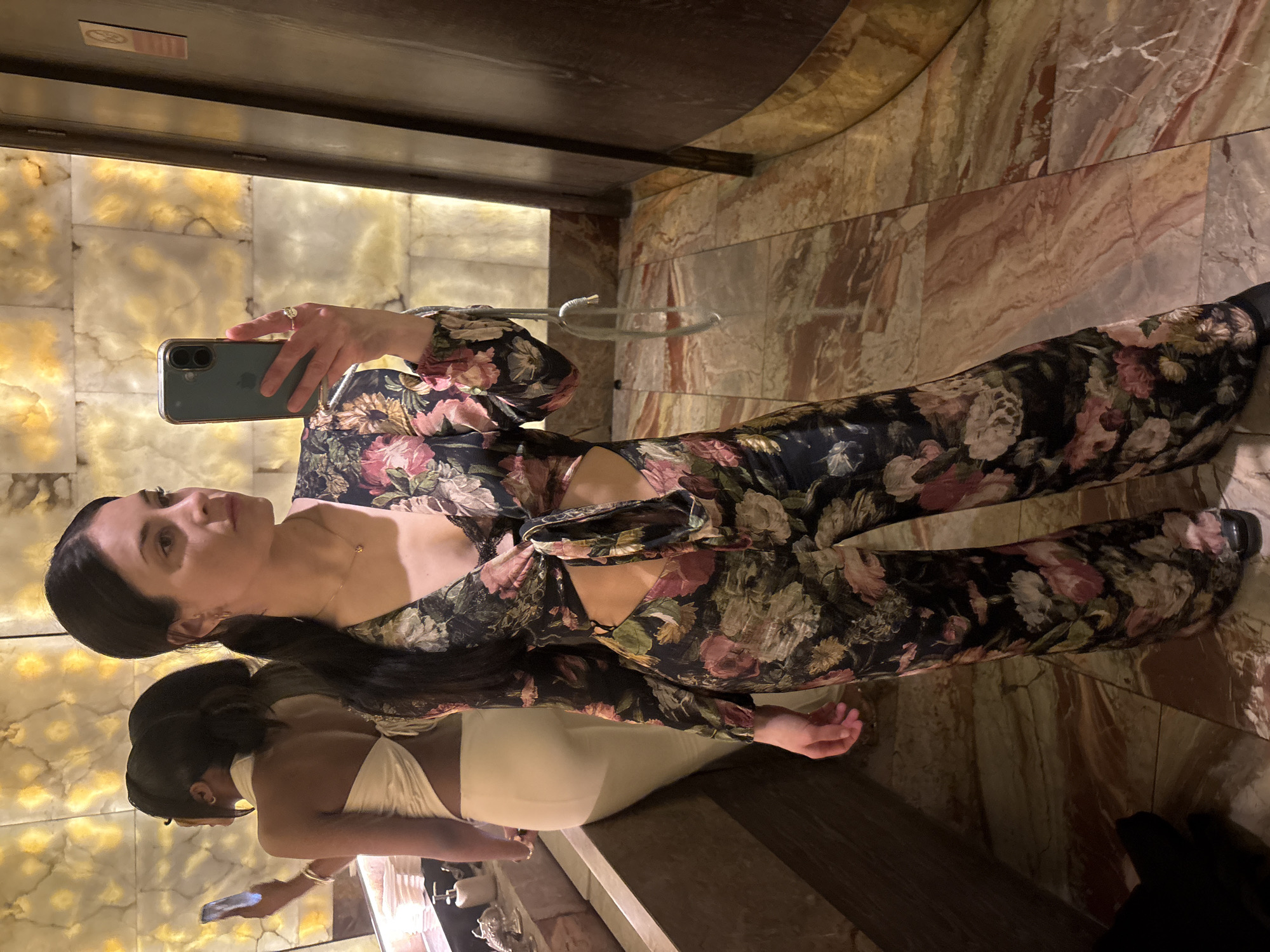 A woman with long dark hair takes a selfie in a bathroom. She is wearing a floral-patterned jumpsuit with cutouts. In the reflection of the mirror, another woman wearing a light-colored outfit stands nearby. The bathroom features marble floors and walls. The location is likely within a building in London, United Kingdom, based on the style of the interior design.