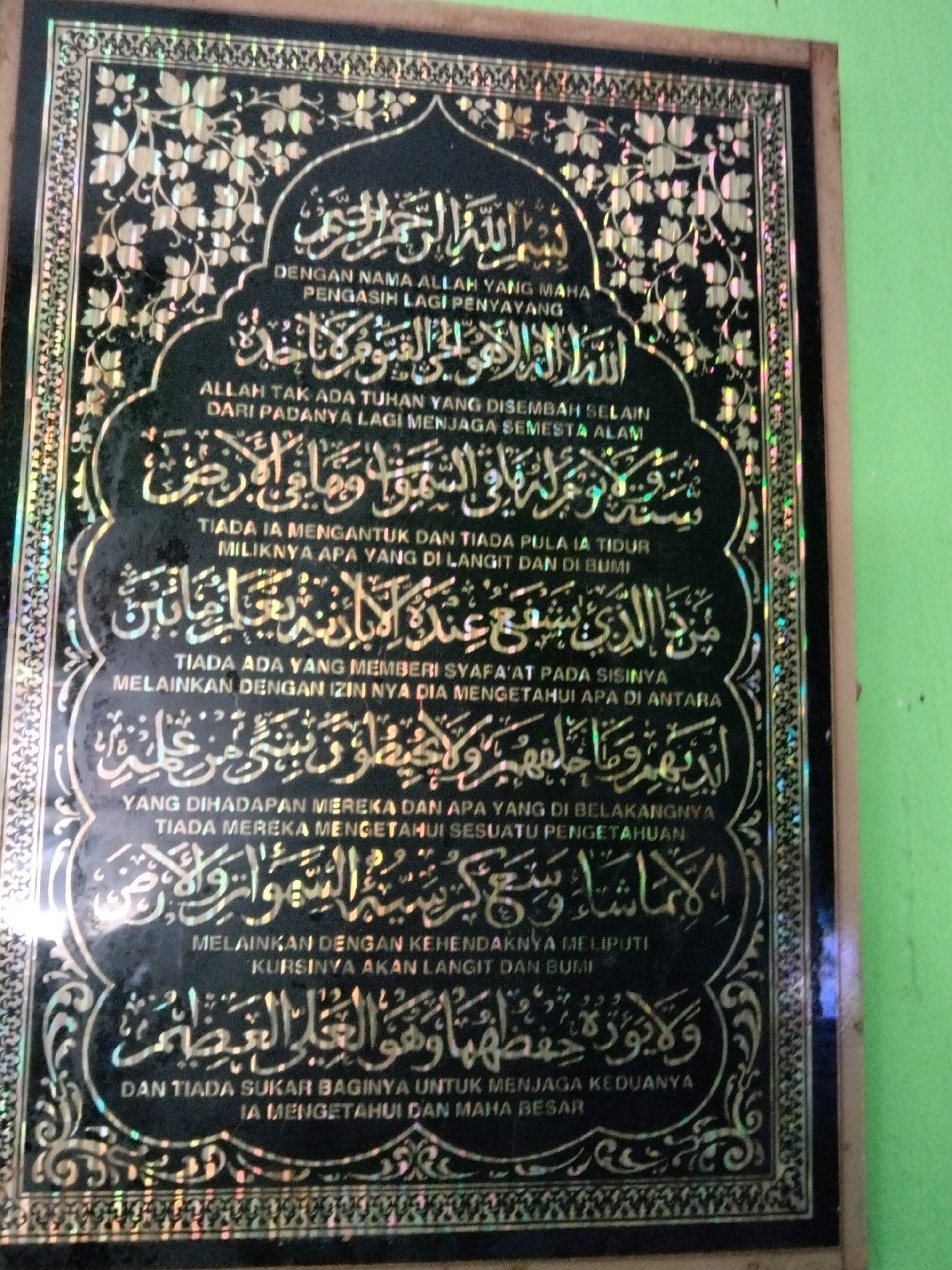 A framed Islamic calligraphy artwork hangs on a light green wall. The artwork is black with intricate gold floral patterns and decorative borders. The text, also in gold, is in Arabic script and appears to be a religious verse. The framed artwork is centrally positioned on the wall. To the left of the artwork, a sliver of a dark object, possibly a wooden frame or another wall section, is visible. The lighting in the scene appears to be artificial, casting a slight glare on the artwork. The overall environment suggests an indoor setting, likely a home or a place of worship. There are no people or animals present in the image.