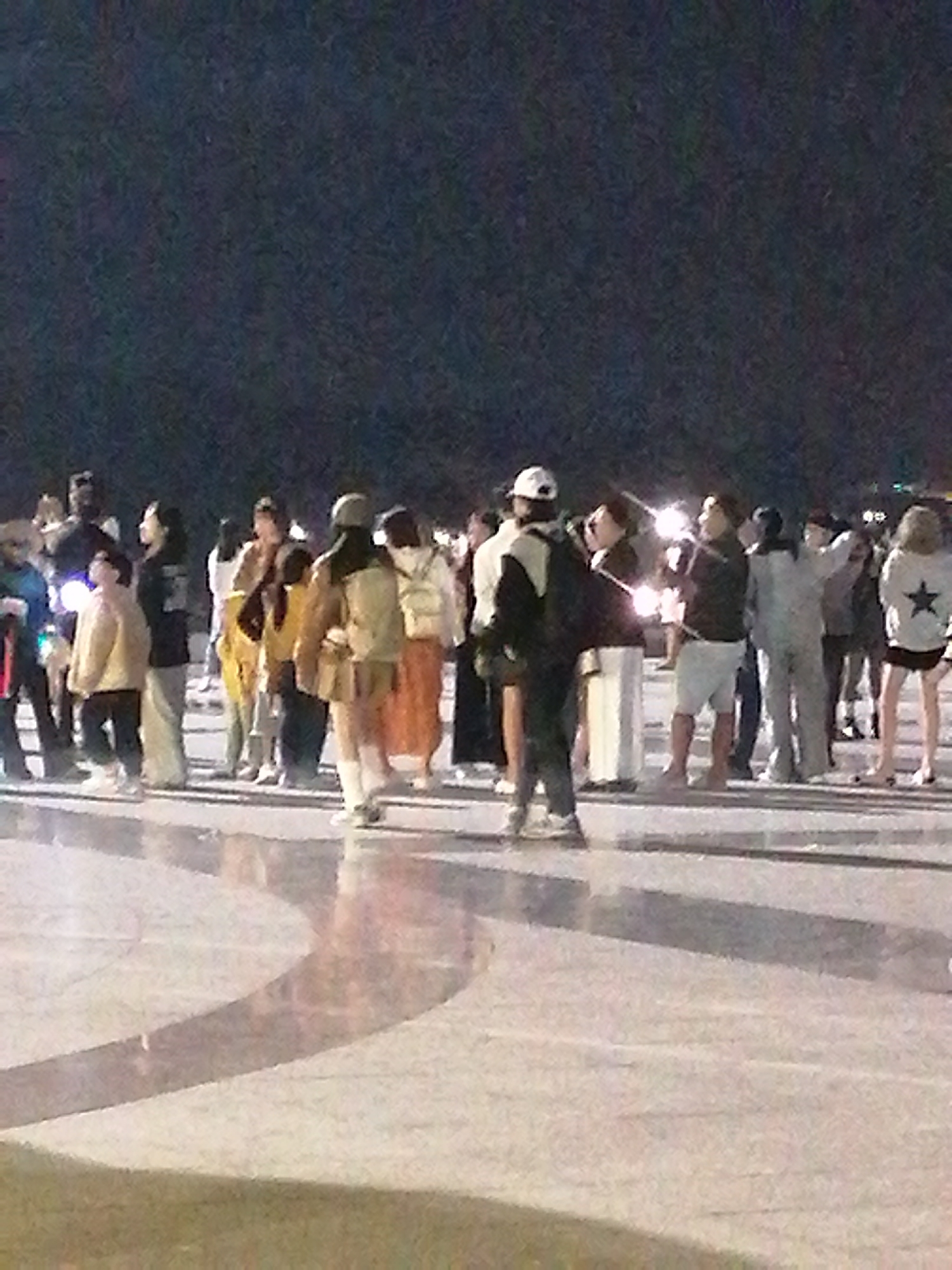 In the city of Da Lat, Vietnam, a group of people stand on a paved surface at night. The ground is a light-colored stone with curved patterns, suggesting a public plaza or promenade. The background is dark, indicating nighttime, with some indistinct lights visible. The individuals are diverse in age and attire, some wearing jackets and casual clothing. The focus is on the gathering of people, with some appearing to be interacting or observing something. The scene is lit by artificial lights, casting subtle shadows.
