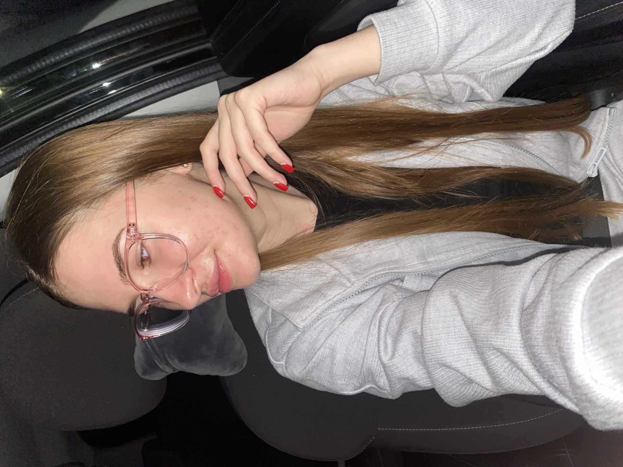 A young woman with long brown hair and pink-rimmed glasses is pictured in the interior of a vehicle. She is wearing a light gray zip-up jacket over a black shirt. Her right hand is resting on her chin, and her fingernails are painted red. The background shows the dark interior of a car, including a headrest and part of a door. There is no visible indication of the specific location within Дьяково, Russia.