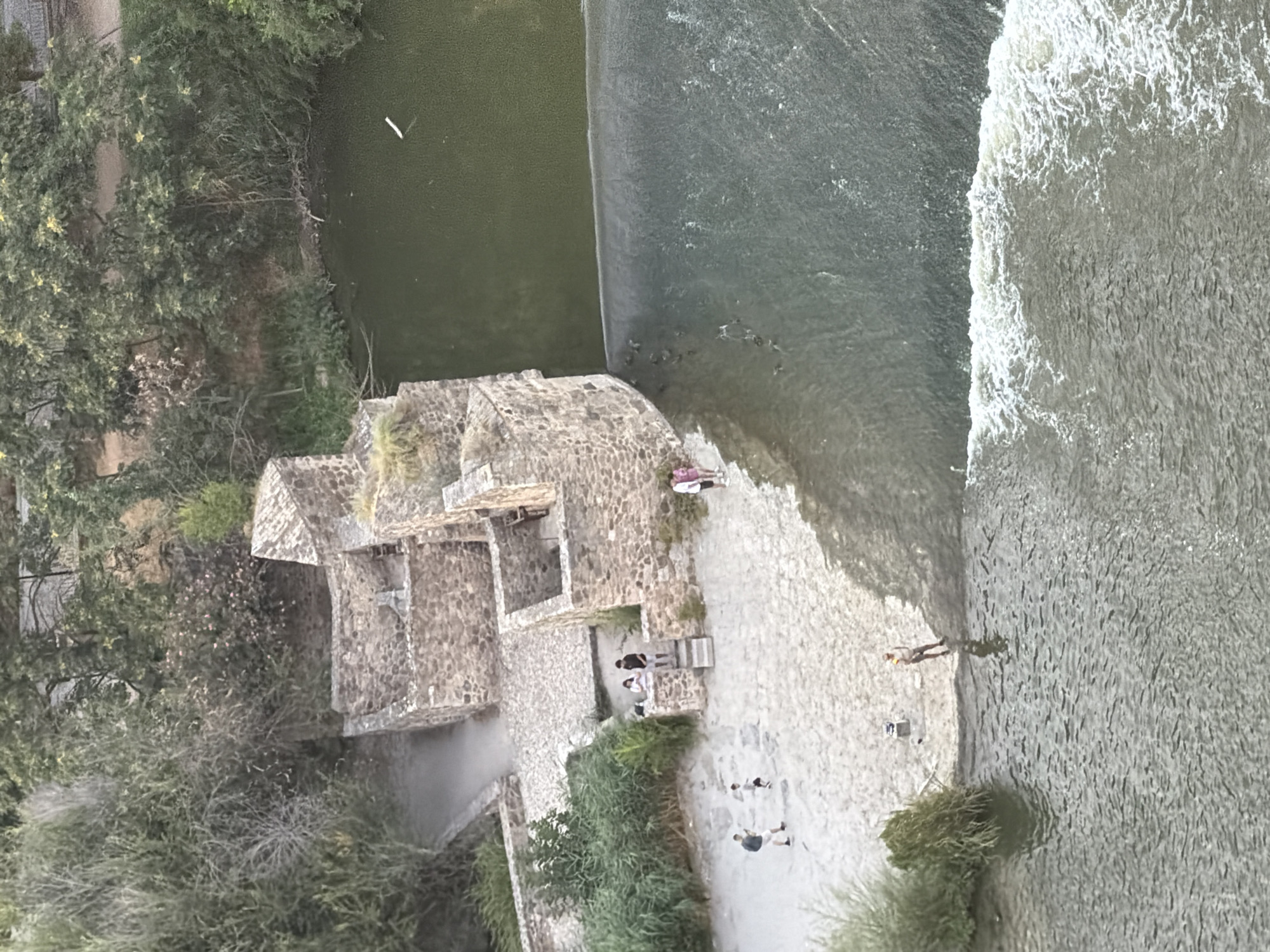 The image captures a high-angle view of the Tagus River in Toledo, Spain, focusing on a historical stone structure situated along its bank. The structure appears to be a ruin, with several stone walls forming distinct sections and staircases. Lush green vegetation surrounds the ruins and the riverbank.
In the foreground, the river flows with a visible waterfall or weir creating white rapids at the edge. The water is a murky green. On the sandy bank adjacent to the weir, several individuals are present. A man in a light-colored shirt and shorts stands near the water's edge, possibly fishing or observing. Further up the bank, a group of people, including two adults and a child, are gathered near a set of stone steps leading up to the ruins. Two women in casual clothing are also visible on the sandy bank.
The ruins themselves are constructed from rough-hewn stones, with openings suggesting doorways and windows. The surrounding environment is a mix of natural foliage and the stone architecture, suggesting a historical site integrated into the natural landscape. The overall scene depicts a peaceful daytime setting at a scenic river location.