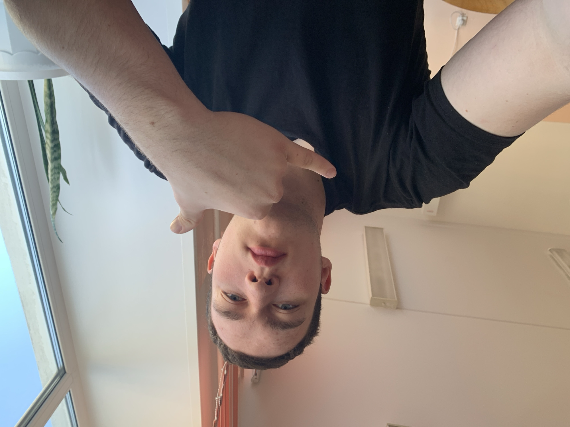 A young, fair-skinned individual is seen in a selfie taken at an inverted angle. He is wearing a black t-shirt and making a hand gesture toward himself. The background features a window with a view of a blue sky and a green plant. The room also has white walls with electrical outlets. The location cannot be determined as Severodvinsk, Russia, based on the provided image.