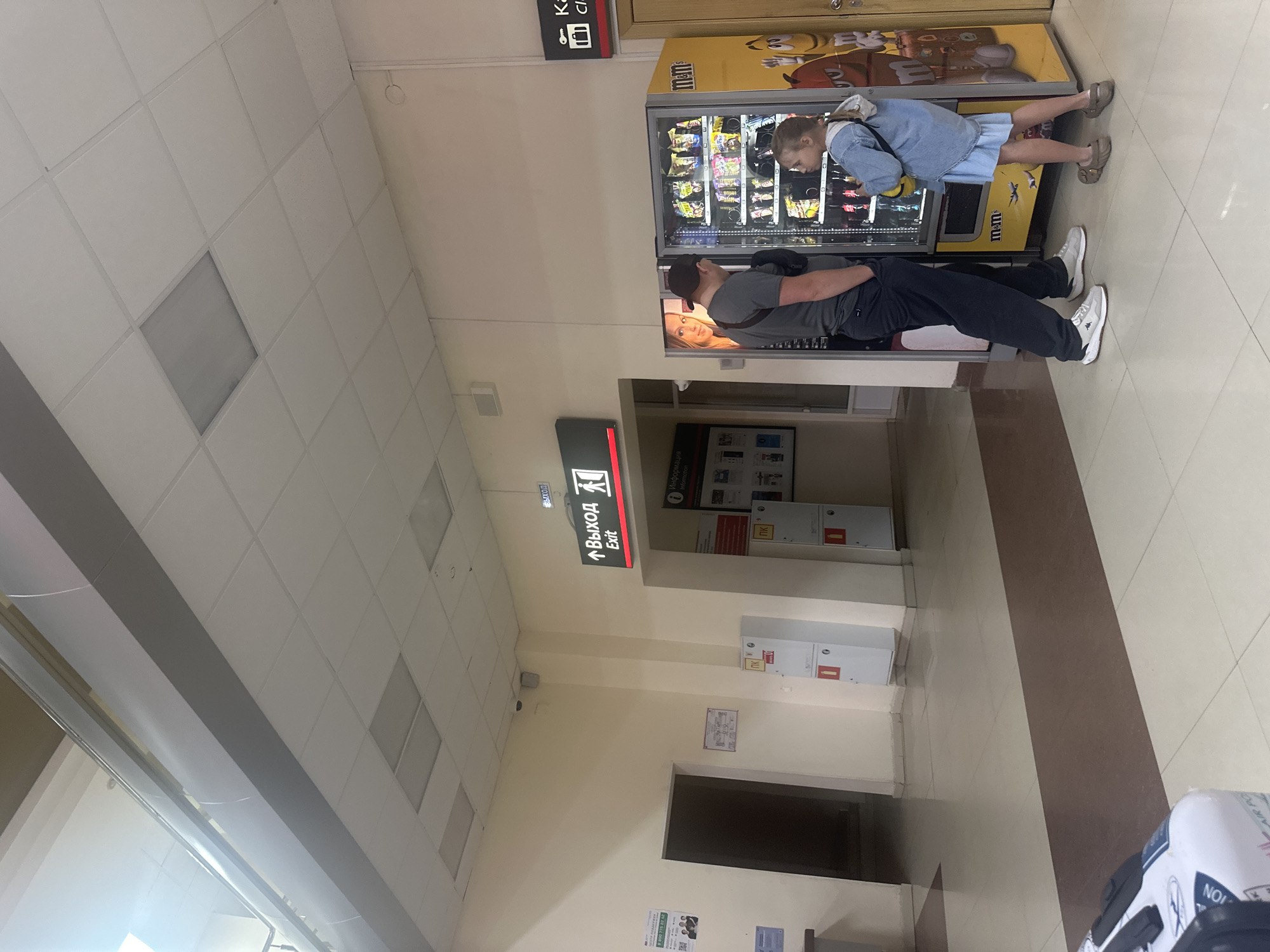 In Lyambir', Russia, a young girl in a light blue denim dress and jacket is reaching into an M&M's vending machine. She appears to be contemplating her purchase. Beside her, a man in a dark grey t-shirt and black pants, wearing a black baseball cap, stands with his arm around her, also looking at the machine. He has his eyes closed, and his head is tilted slightly, suggesting a moment of quiet intimacy or shared enjoyment.
The setting appears to be an indoor public space, possibly a shopping mall, airport, or train station, indicated by the tiled floor, suspended ceiling tiles, and illuminated exit signs in both Russian and English. One sign clearly reads "Выход Exit". Other signs and fixtures suggest a commercial or service area, including information boards and what looks like other vending machines or service points. The lighting is bright and artificial, suggesting it could be daytime or evening. There are no specific indications of the weather. The overall mood seems relaxed and casual, focused on the interaction between the two individuals and the vending machine.