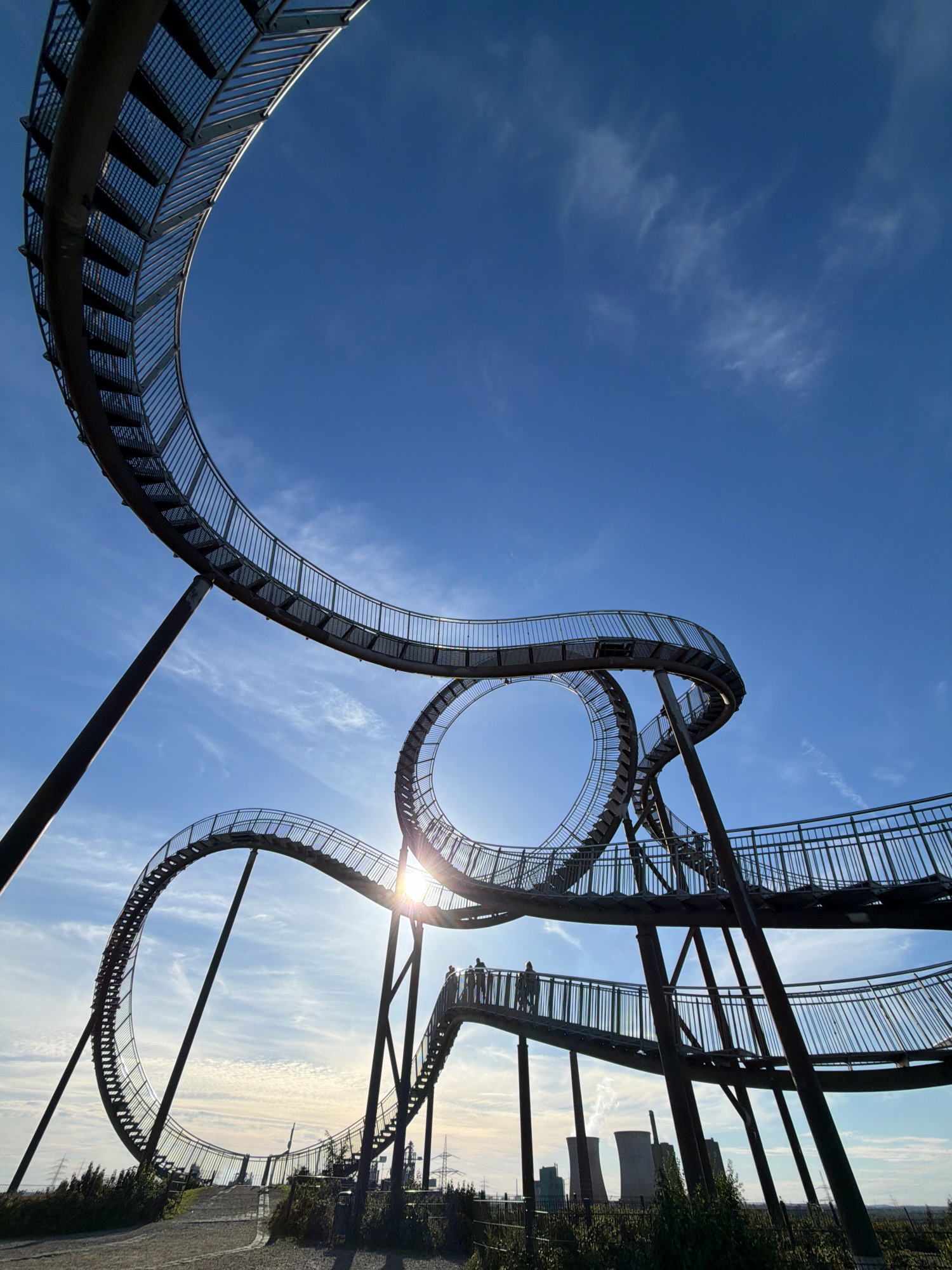 The image depicts the "Tiger and Turtle – Magic Mountain" sculpture, an art installation located in the city of Duisburg, Germany. The sculpture, which resembles a roller coaster, is made of steel and features a winding pathway with stairs and ramps. It is set against a bright blue sky with scattered white clouds. Several people can be seen walking on the sculpture, enjoying the panoramic views. In the background, industrial cooling towers are visible, hinting at the location's industrial heritage. The sun is shining, casting a warm glow on the scene.