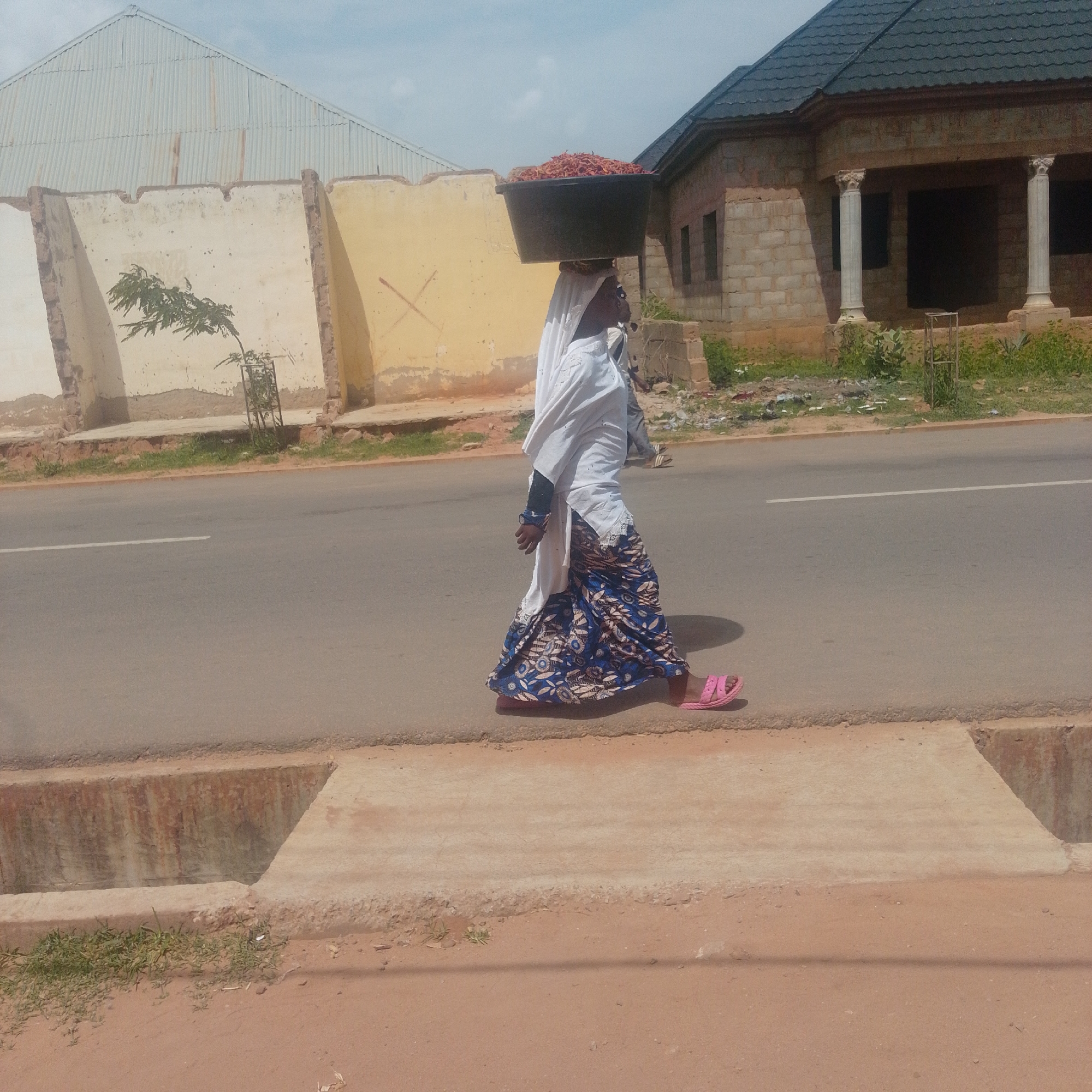 A woman walks along a paved road in Gombe, Nigeria, carrying a large black basin filled with red chili peppers on her head. She is dressed in a white hijab and a blue and white patterned skirt, and she wears pink sandals. The woman is walking on the side of the road, next to a drainage ditch. In the background, there are buildings under construction, one with a dark tiled roof and columns, and another with a corrugated metal roof. The sky is partly cloudy, suggesting it is daytime. The scene appears to be a typical street in a populated area, possibly a marketplace or residential neighborhood, with the woman engaged in the activity of transporting goods for sale or personal use.
