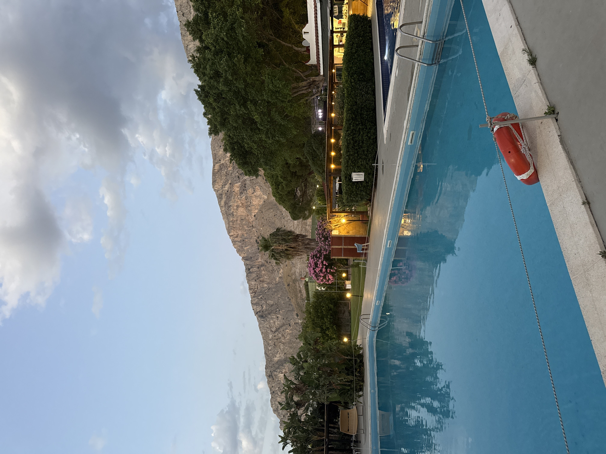 The image depicts a serene outdoor swimming pool area at dusk, with a dramatic mountain range serving as a backdrop. The pool is filled with clear blue water, reflecting the sky and surrounding greenery. Along the edge of the pool, a red life preserver is attached to a metal post. A short distance away, a smaller pool with steps and a railing is visible. The area is landscaped with lush trees, bushes, and flowering plants, creating a vibrant and inviting atmosphere. In the background, a building with outdoor lighting suggests a bar or restaurant area, and beyond that, the imposing mountain looms against a sky filled with soft clouds. The scene appears to be located in Isola delle Femmine, Italy, a coastal town known for its natural beauty and proximity to Palermo.