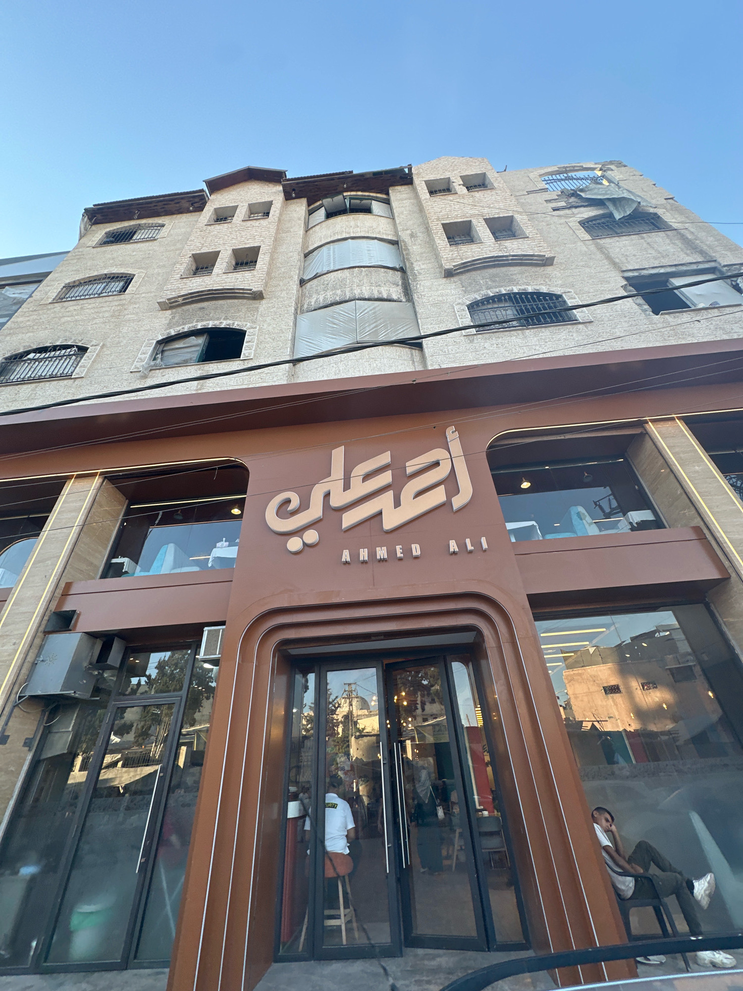 The image depicts the facade of a multi-story building in the city of Gaza, Palestinian Territories. The building's upper floors show signs of damage, with some windows broken or covered with plastic sheeting, and parts of the roof appearing dislodged. Below these damaged floors, the lower section of the building is a modern establishment with a brown facade featuring signage in Arabic script and English lettering that reads "AHMED ALI". The lower level has large glass windows and a prominent entrance. Inside the establishment, visible through the windows, are tables and seating, suggesting it is a commercial space, possibly a restaurant or cafe. People are visible inside and outside the building, including individuals seated at tables and a person in a white shirt with the word "SECURITY" on the back, seated near the entrance. Reflections in the glass show other buildings and the sky.