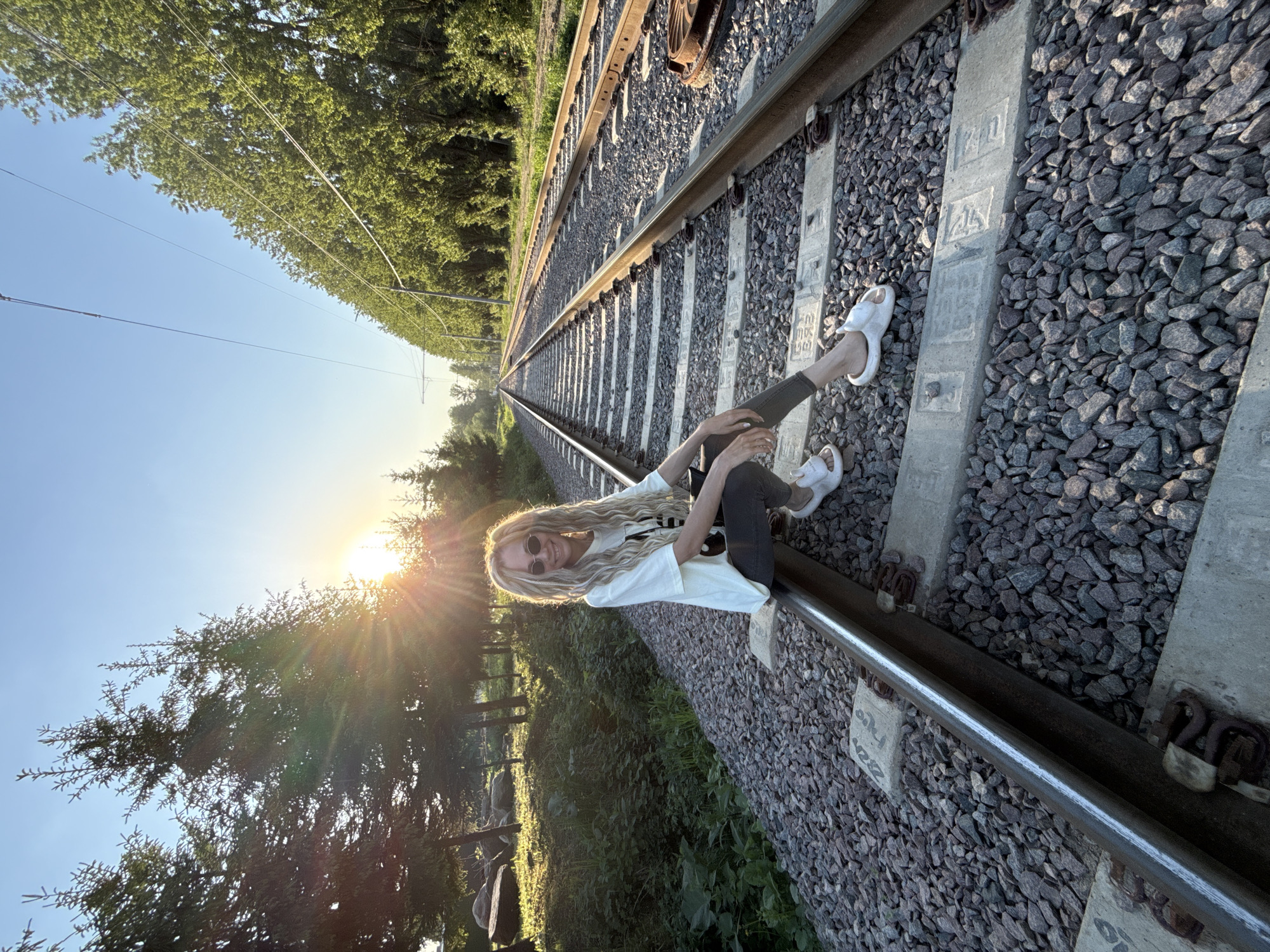 A long-haired woman wearing sunglasses, a white t-shirt, dark pants, and white sandals is sitting on the railway tracks. The railway tracks extend into the distance, with gravel and concrete sleepers visible. On either side of the tracks, trees and vegetation are present. The sun is setting, casting a bright glare in the background. The photo was taken in Заволжье, Russia.