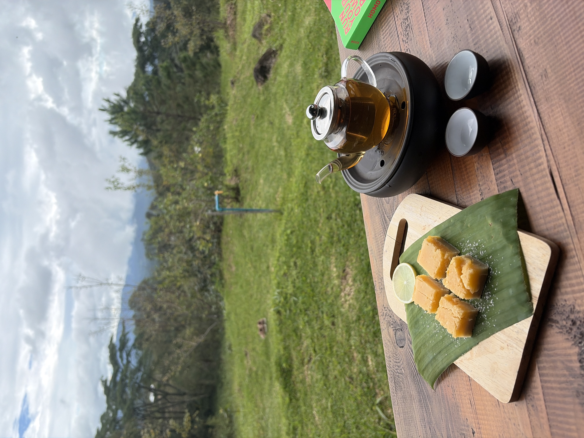 The image shows a tea set and some snacks placed on a wooden table outdoors, with a view of a lush green landscape under a cloudy sky. The scene appears to be set in Ban Khun Wang, Thailand, given the surrounding vegetation which is typical of the region. In the foreground, a clear glass teapot filled with amber-colored tea sits on a black circular stand. Next to it are two small, dark-rimmed ceramic cups. To the left, on a wooden cutting board topped with a banana leaf, are four square pieces of light orange confectionery, possibly fudge or a similar sweet, dusted with white powder. A slice of lemon and a small cinnamon stick rest on the banana leaf. In the background, the landscape features rolling hills covered in trees, with a visible pathway or clearing. The sky is overcast with large, fluffy clouds, suggesting a potentially cool or humid day. The overall atmosphere is one of a tranquil, outdoor tea-time experience.