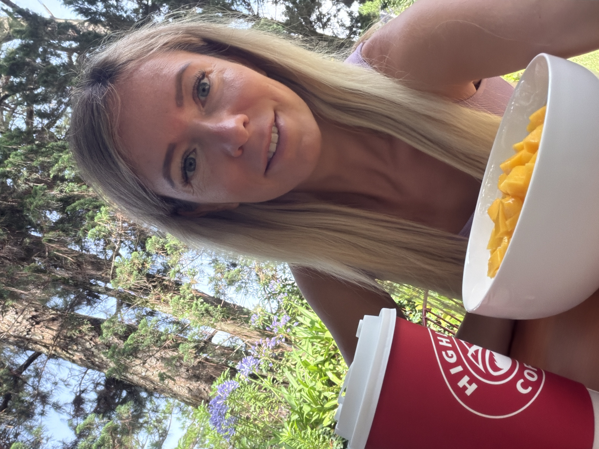 A blonde woman is taking a selfie outdoors, with a cup of coffee and a bowl of fruit in the foreground. She is smiling and looking at the camera. The background consists of trees and some purple flowers. The setting appears to be a garden or a park.