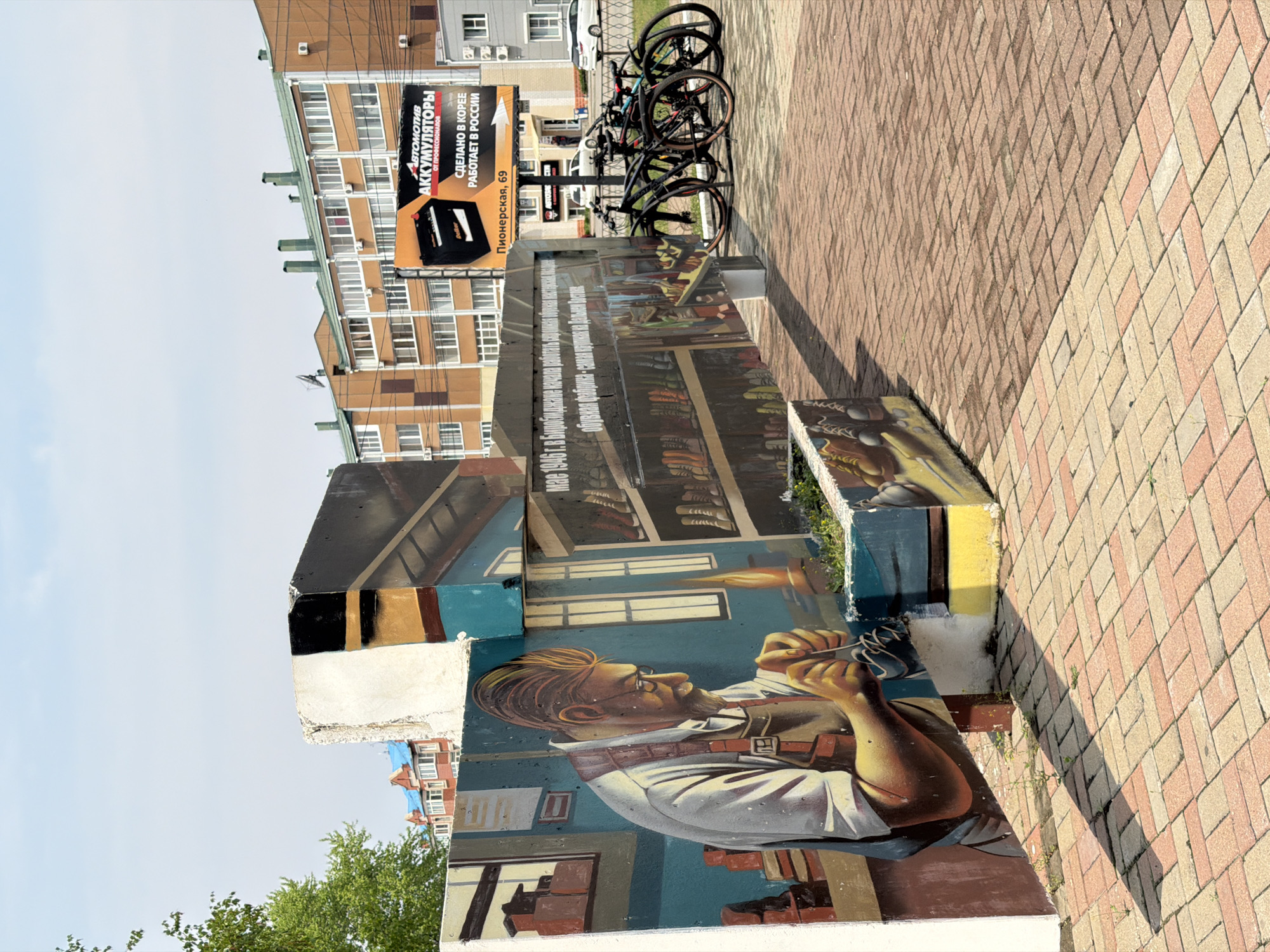 The image shows a painted mural on a structure in Birobidzhan, Russia. The mural depicts a bespectacled man in a shirt with suspenders holding tools, likely a craftsman or artist. Above him is another section of the mural with shelves of books and what appears to be a fiery object. Adjacent to the mural, there is a sign for "АВТОМОТИВ АККУМУЛЯТОРЫ" (Automotive Batteries) that says "СДЕЛАНО В КОРЕЕ РАБОТАЕТ В РОССИИ" (Made in Korea, Works in Russia). Several bicycles are parked behind the mural. The mural and sign are located on a brick-paved sidewalk. A multistory apartment building is visible in the background. The sign is located on Pionerskaya Street at number 69.