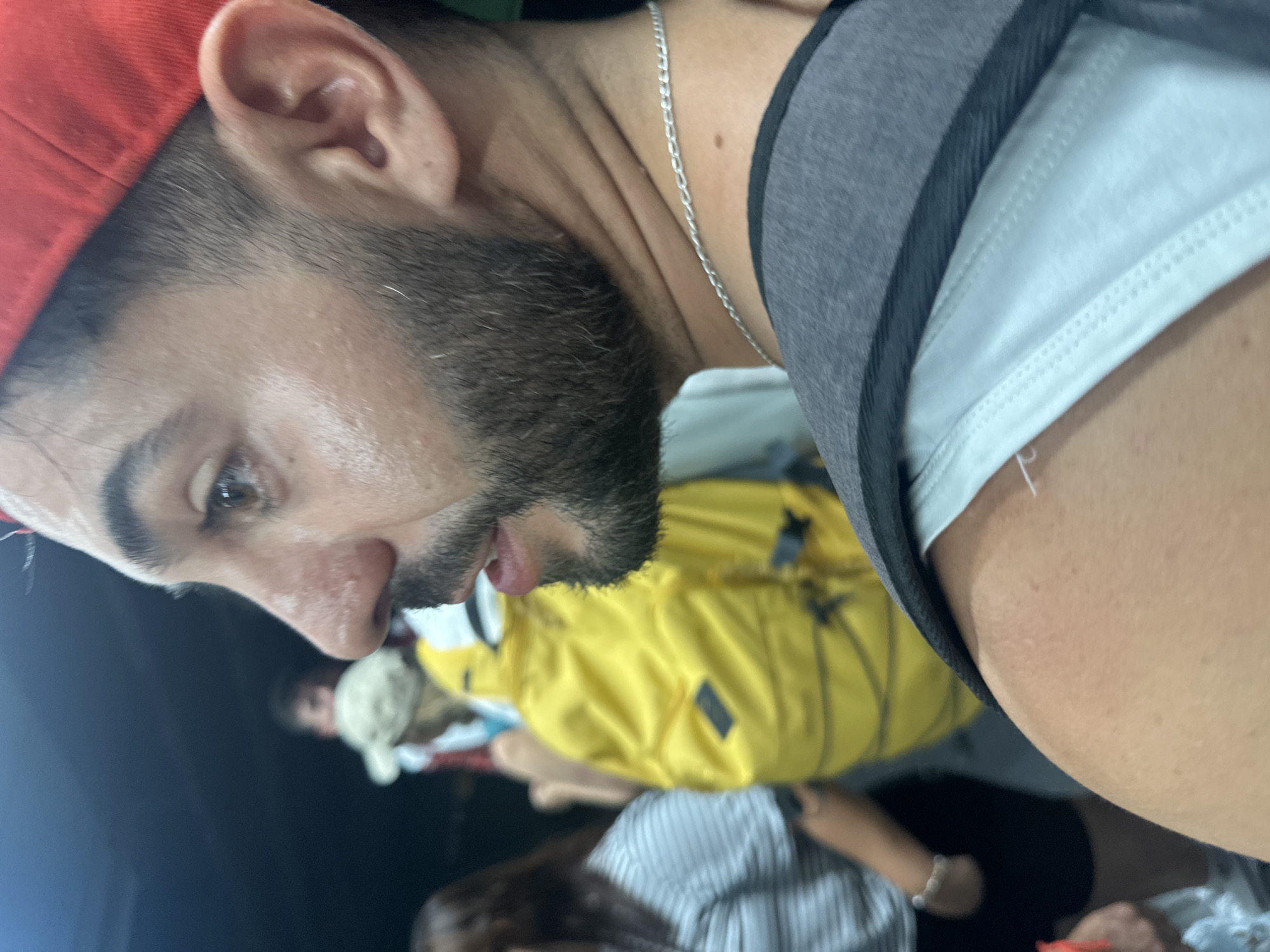 The image shows a medium shot of a man wearing a red baseball cap and a silver chain. He has a beard and is looking to the side. In the background, other people are visible, one wearing a yellow jacket and another in a striped shirt. The environment appears to be outdoors, possibly a crowded public space, with dim lighting. There is no discernible indication of the specific location within Barcelona, Spain.