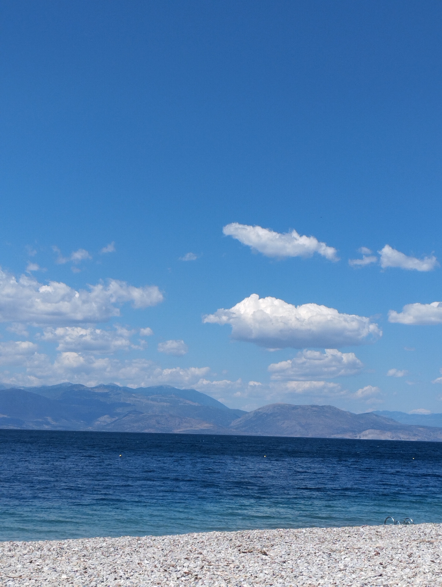 This is a landscape photograph of a tranquil beach scene. The foreground is dominated by a wide expanse of small, smooth pebbles, likely a pebble beach. Beyond the pebbles, a body of deep blue water stretches towards a distant, hazy mountain range. The sea is calm, with gentle ripples visible on its surface, transitioning to a lighter turquoise hue closer to the shore. The sky above is a clear, vibrant blue, punctuated by scattered, fluffy white clouds. The light suggests it is daytime, possibly mid-morning or early afternoon, with the sun illuminating the scene evenly. There are no people or discernible objects in the immediate foreground or mid-ground, emphasizing the natural beauty and serenity of the location. The mountains in the distance appear rugged and undeveloped, suggesting a natural, possibly remote, setting. The overall mood is peaceful and inviting, evoking a sense of calm and relaxation. There is no visible text in the image.