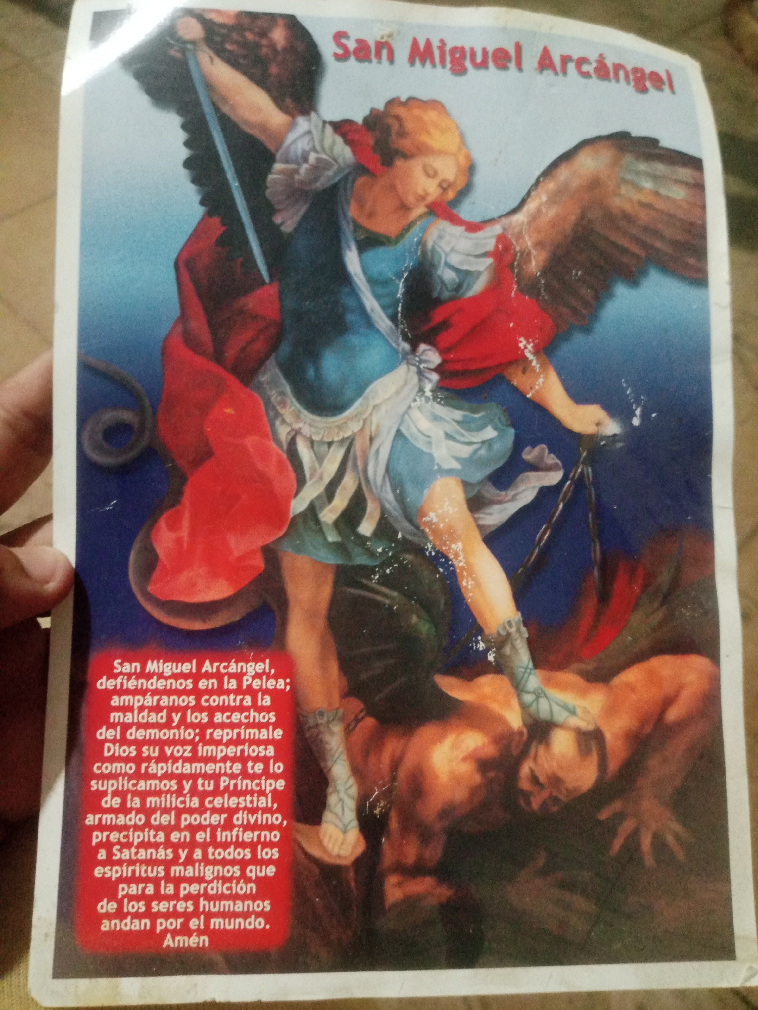 The image depicts a religious print featuring Saint Michael the Archangel, identified by the Spanish text "San Miguel Arcángel." He is portrayed in a dynamic pose, with wings spread, holding a sword aloft and stepping on a fallen figure, presumably Satan. Saint Michael is dressed in a blue tunic, red cape, and armored greaves, while the fallen figure is rendered in earthy tones. The background is a gradient of blue, with some red elements suggesting fire or hell. Below the image, a prayer to Saint Michael is inscribed in Spanish. This print is a devotional item, commonly found in Catholic households and churches. The image does not provide any specific information about its location in Santa Ana, Venezuela, or any urban context.
