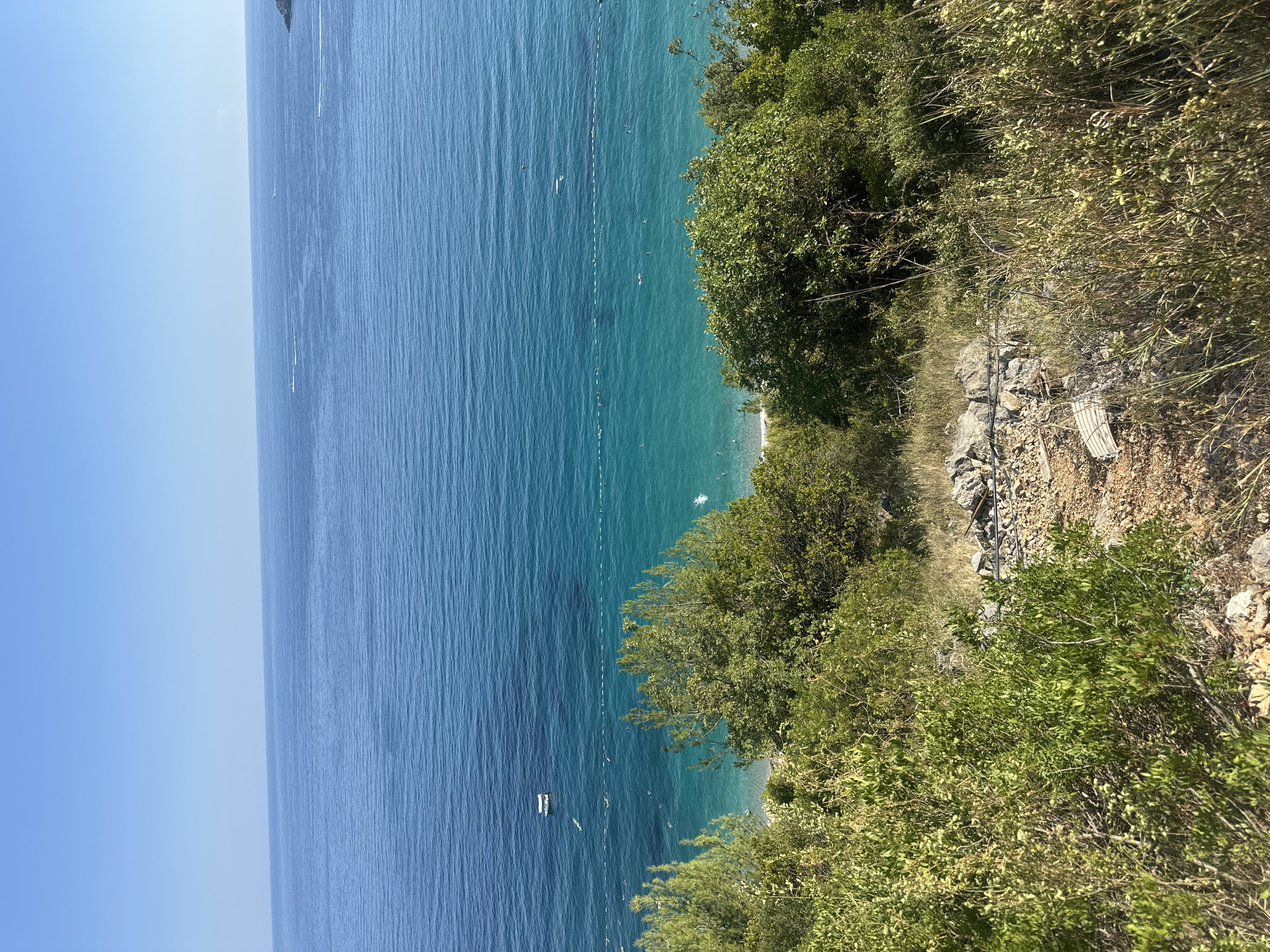 The image captures a scenic view of the Adriatic Sea from a hillside location near Prijevor, Montenegro. In the foreground, lush green vegetation and rocky terrain dominate, with dry grasses and scattered stones. Beyond this, the turquoise waters of the sea stretch out to the horizon under a clear blue sky. A beach area is visible along the coastline, with a line of buoys marking a swimming area. Several small boats are present on the water, including a white motorboat closer to shore and two speedboats further out. A few individuals can be seen swimming in the sea. The overall impression is one of a sunny, tranquil coastal landscape.