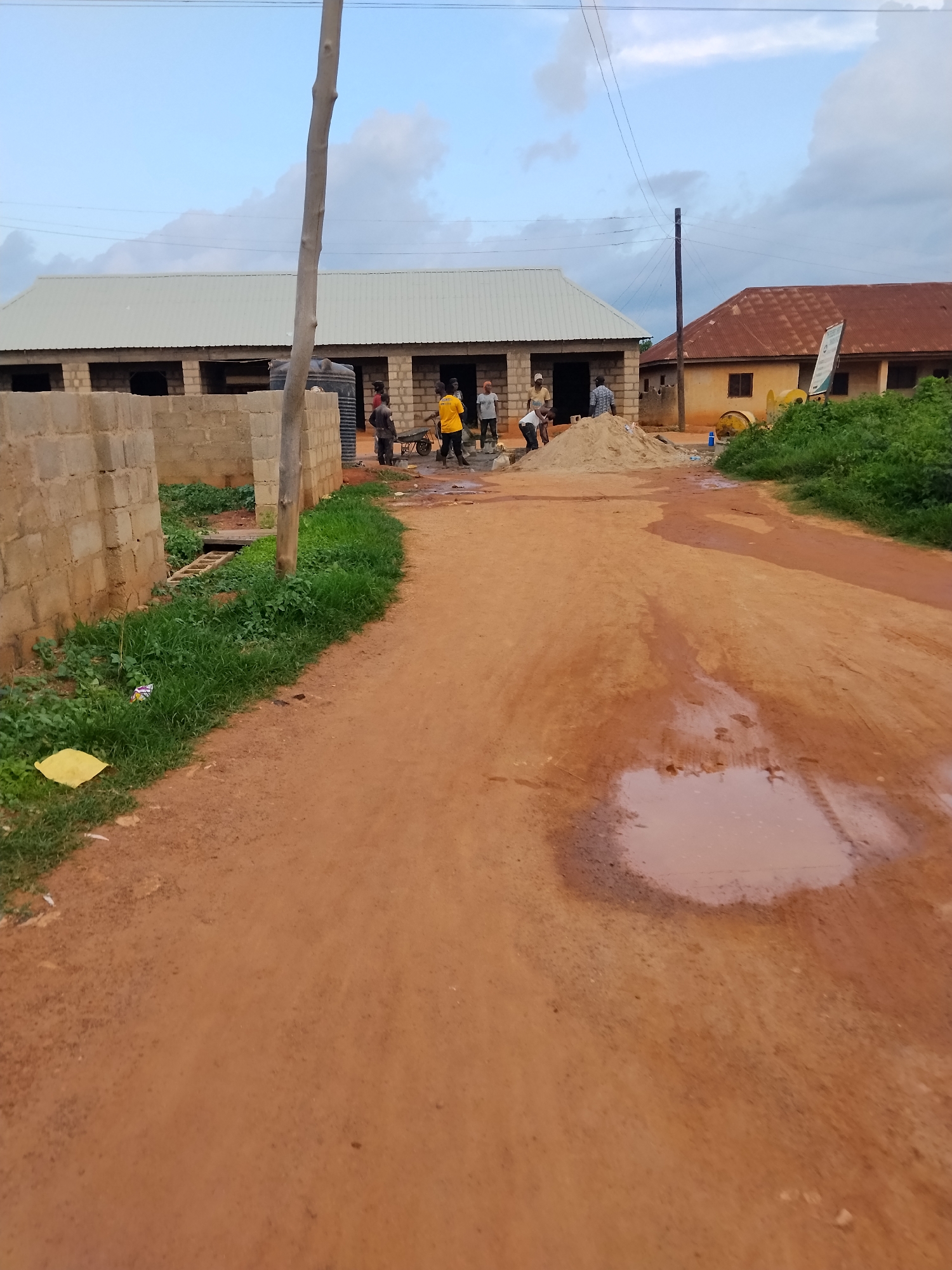 The image depicts a construction site in Mangu Town, Nigeria, with several individuals engaged in work. In the foreground, a dirt road occupies the majority of the frame, with a large puddle of muddy water reflecting the sky. To the left of the road, a partially constructed building made of concrete blocks is visible, with a water tank nearby. Several people are working on this structure, some carrying materials and others appearing to be mixing cement. Further into the scene, another building with a corrugated metal roof is under construction. In the background, a second building with a reddish-brown metal roof stands, and a yellow cement mixer is present. Lush green vegetation borders the right side of the frame. Power lines are strung across the sky, supported by a wooden pole and a utility pole. The overall impression is one of ongoing construction work in a semi-urban environment.
