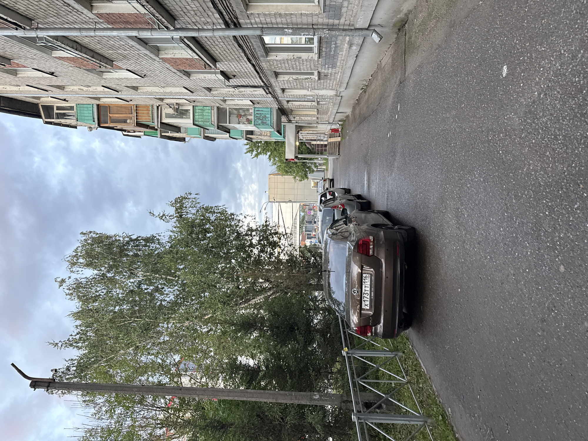 This is a medium-shot, eye-level, outdoor photograph taken on a cloudy day in Kazan, Russia. The scene depicts a paved walkway or street running alongside a multi-story brick apartment building. Several cars are parked along the walkway. The closest car, a brown Volkswagen sedan, is facing away from the camera, its license plate clearly visible as "X173TT 116". To the left of the walkway is a metal fence and a grassy area with trees. A concrete utility pole stands near the fence, topped with a light fixture. The apartment building is light gray brick, with several balconies visible, some painted green and others wooden. The sky is overcast with dramatic cloud formations.
