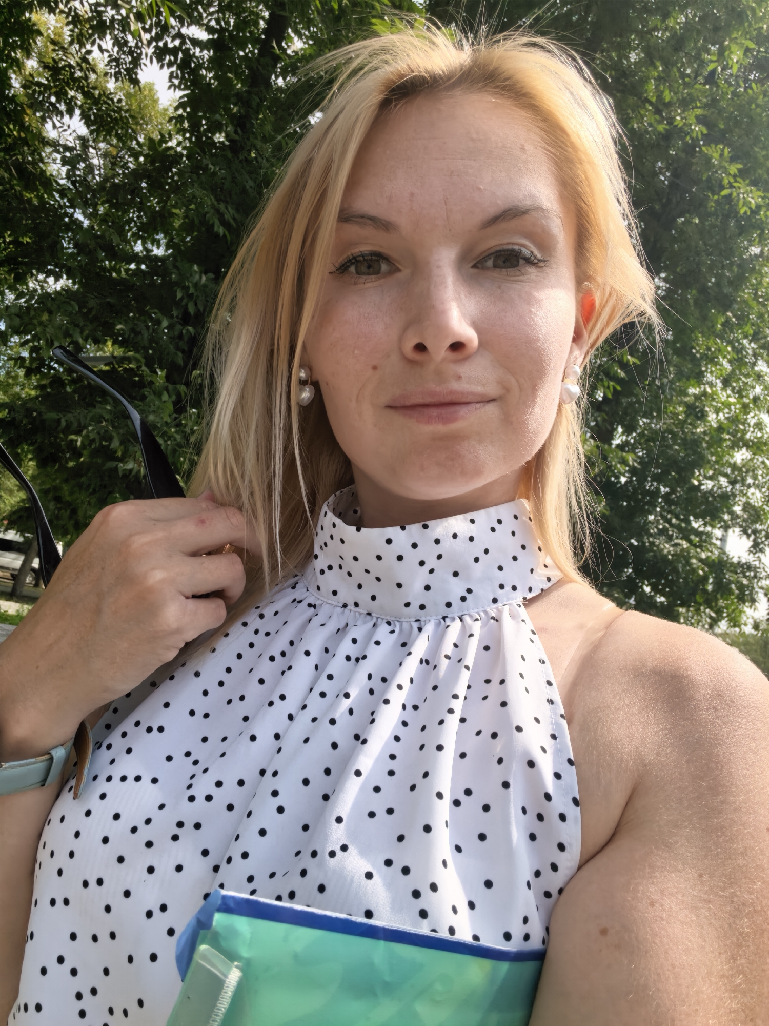 A blonde woman with fair skin, wearing a white polka dot halter top and pearl earrings, is captured in a close-up selfie. She is outdoors with lush green trees and foliage in the background, suggesting a park or a natural setting. The lighting indicates it is daytime, possibly sunny. Her left hand is visible, holding a pair of dark sunglasses. A light blue watch is on her left wrist, and she is holding a teal and blue object, possibly a bag or a folder, in front of her. The image does not provide any specific indicators of the city of Sterlitamak, Russia.