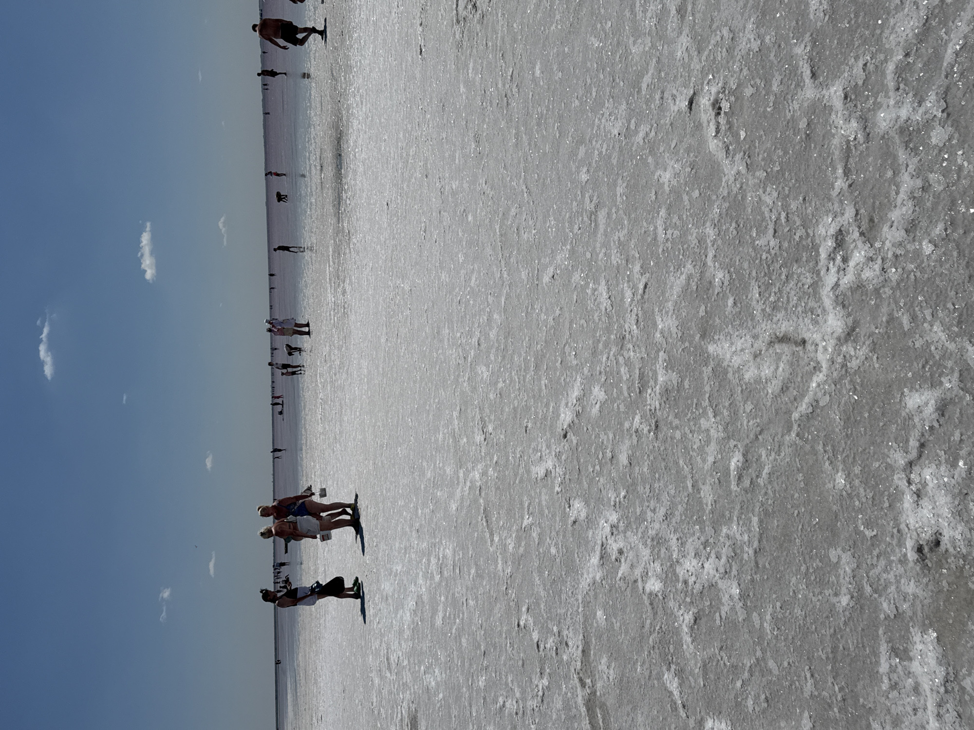 A group of people are visible on the pink salt flats of Bursol', Russia, under a clear blue sky with scattered white clouds. The foreground of the image is dominated by the textured surface of the salt flats, with a crystalline, uneven appearance. In the middle ground, several individuals are scattered across the expanse. Three women stand together near the left side of the frame, one holding a black bag. Further back, other figures can be seen walking or standing, some appearing to be collecting something from the salt flats. A man is walking away from the camera on the right side of the frame. The horizon line is visible in the distance, where the pink water of the salt lake meets the sky.