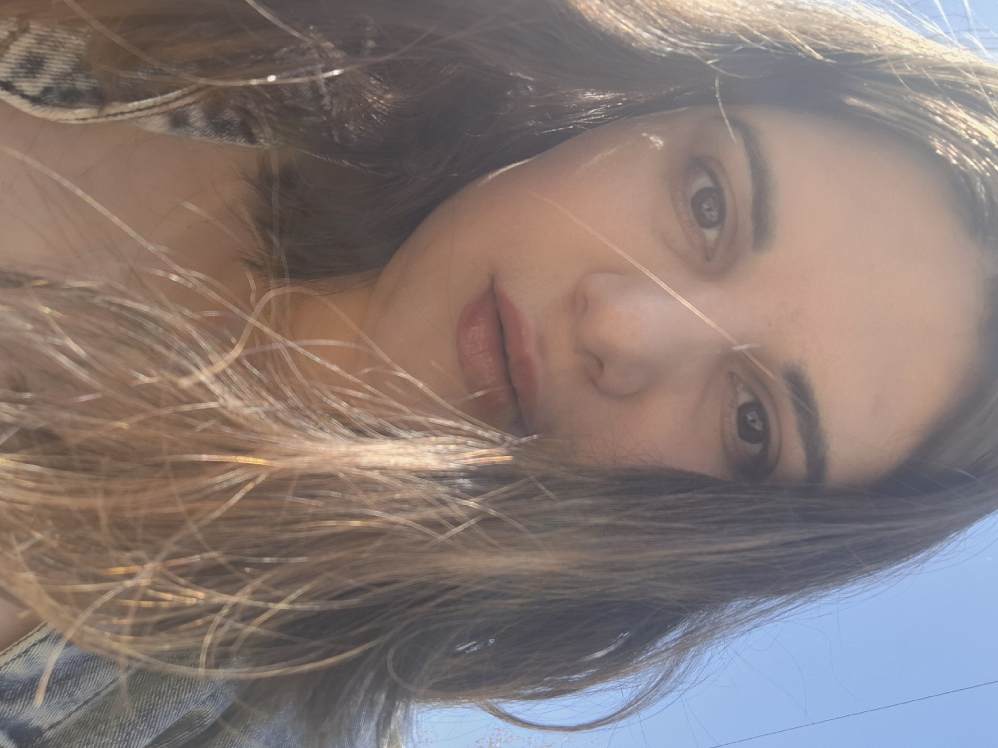 A medium close-up, tilted shot captures a woman with long, brown hair partially obscuring her face. Her skin appears fair. Her brown eyes are visible, and she wears a light lipstick. She is wearing a denim jacket. A clear blue sky is visible in the background. The location is Chaikovsky, Russia.