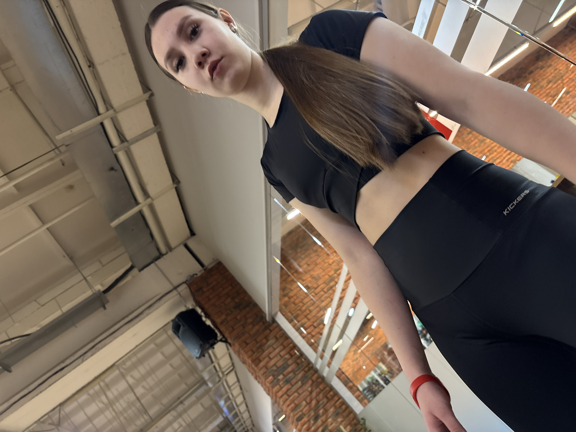 A young woman is captured in an indoor setting, likely a gym or commercial space, in Chaikovsky, Russia. She is the central subject, photographed from a low-angle perspective, looking directly at the camera with a neutral to serious expression. Her dark brown hair is pulled back, and she is dressed in black athletic wear: a short-sleeved crop top revealing her midriff and high-waisted leggings. A red wristband is visible on her left wrist.
The background reveals architectural details, including an industrial-style ceiling with exposed ducts, beams, and overhead lighting fixtures. A large section of a brick wall is visible, both directly and reflected in what appears to be a large mirror or glass partition. A black wall-mounted speaker is also noticeable. The text "KICKERS" is visible on the waistband of her leggings, indicating the brand. The artificial lighting suggests the image could have been taken at any time of day, as there are no natural light sources prominently featured. The overall scene conveys a modern, perhaps active, environment.