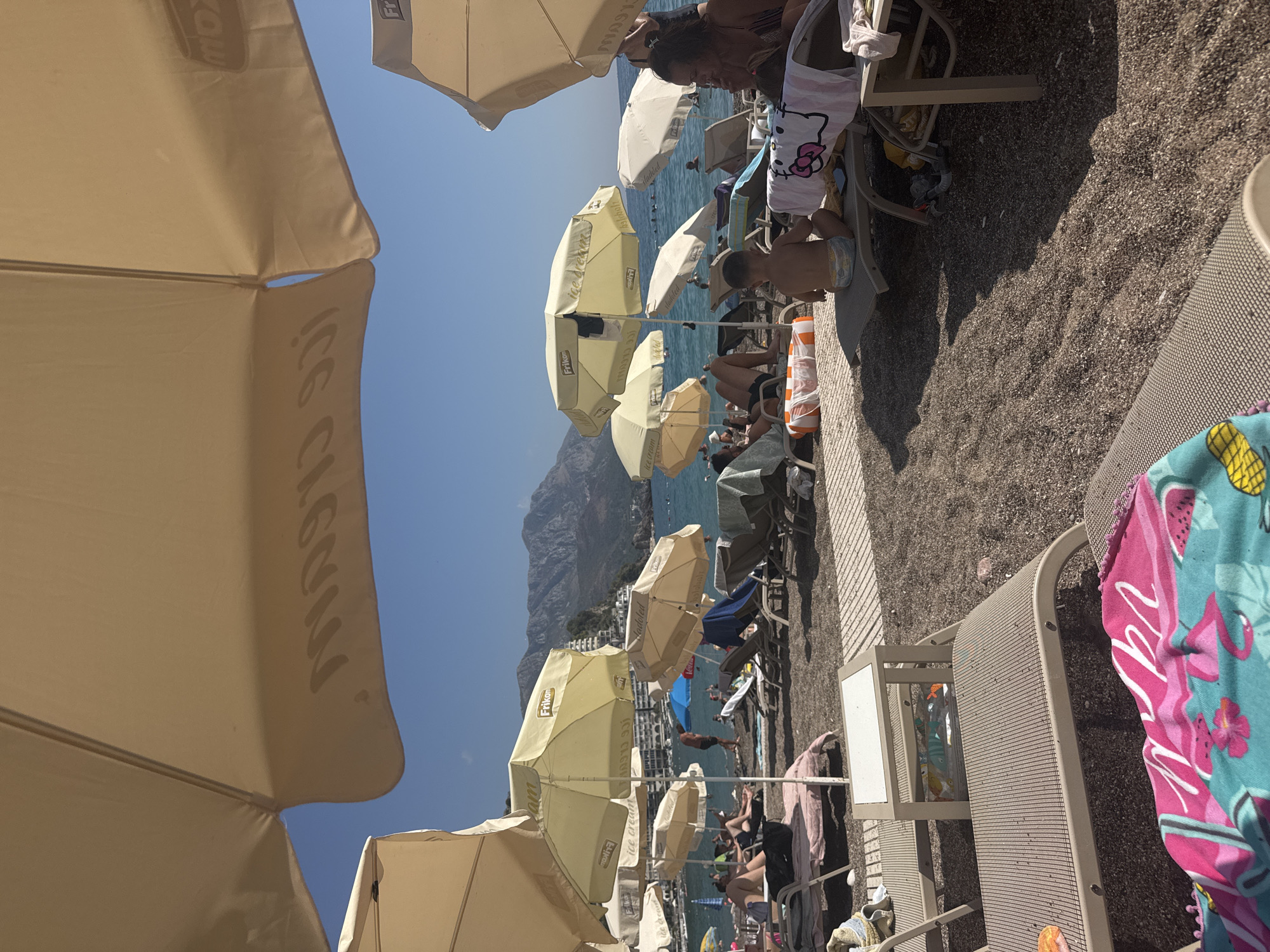 An overhead view of a crowded beach on a sunny day. Numerous umbrellas of various sizes and colors dot the scene, providing shade for beachgoers relaxing on lounge chairs. The beach is covered in dark pebbles, and the sea, with gentle waves, extends to the horizon. In the background, mountains rise, dotted with buildings, and the clear blue sky is visible. People are seen lounging, walking, and interacting on the beach, and some are in the water. A young child sits on a lounge chair in the foreground. Beach accessories like towels and bags are scattered around. The overall impression is a lively and typical summer beach scene.