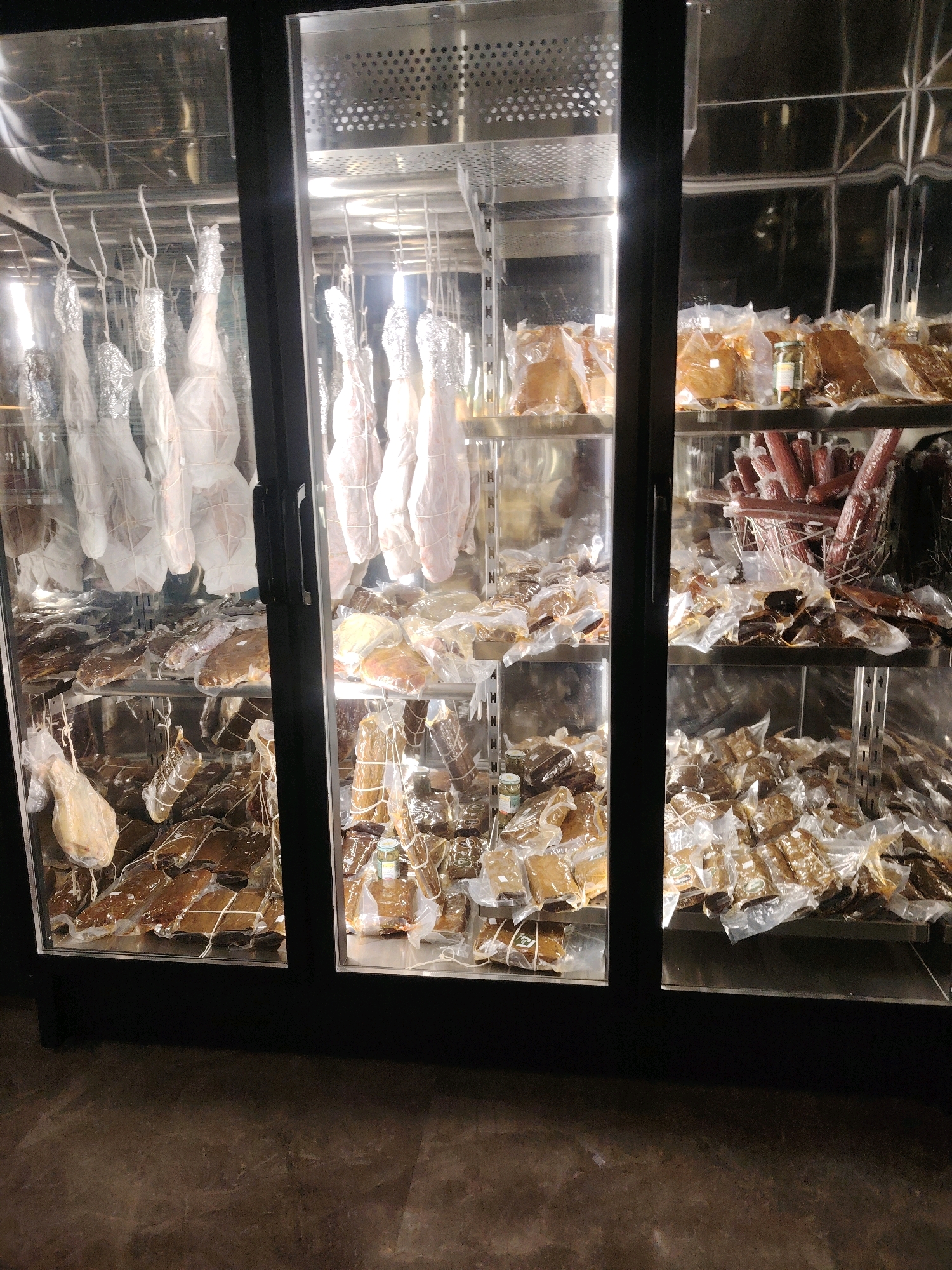 The image depicts a refrigerated display case filled with various cured meats and hams. The display case has three glass doors, and the interior is illuminated, highlighting the products. In the top shelves of the left and center sections, several large hams are hung, wrapped in white cloth and foil. Below these, the shelves are laden with vacuum-sealed packages of cured meats, appearing to be different cuts and types. The right section of the display case also contains vacuum-sealed meats on shelves, with some logs of sausage visible in a basket on one of the upper shelves. The overall environment appears to be a commercial establishment, possibly a deli or market, with a dark tiled floor visible in the foreground. There are no people visible in the image. The location of Konigil, Uzbekistan is not ascertainable from the visual information provided.