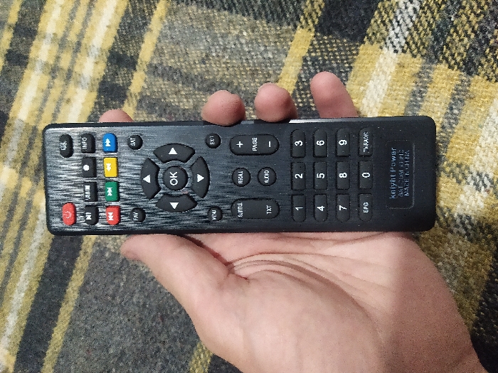 A human hand holds a black remote control, branded "knight Power SAT DVB T2 HE MADE IN CHINA," against a dark grey and yellow striped woven fabric background. The remote, oriented vertically with the top towards the upper edge of the image, features a textured brushed-metal-like finish on its face. Visible buttons include a red power button, several smaller function buttons (e.g., USB, INFO), red, green, yellow, and blue rectangular buttons, a central navigation cluster with an "OK" button and directional arrows, and a numeric keypad (0-9) alongside "MENU," "EXIT," "FAV," "SAT," "TXT," "AUDIO," "RECALL," "SUBTITLE," "EPG," and "TV/RADIO" buttons. The hand's thumb rests near the top-left, with fingers wrapping around the left side of the device. The scene is illuminated evenly. Location: Unknown, Unknown.