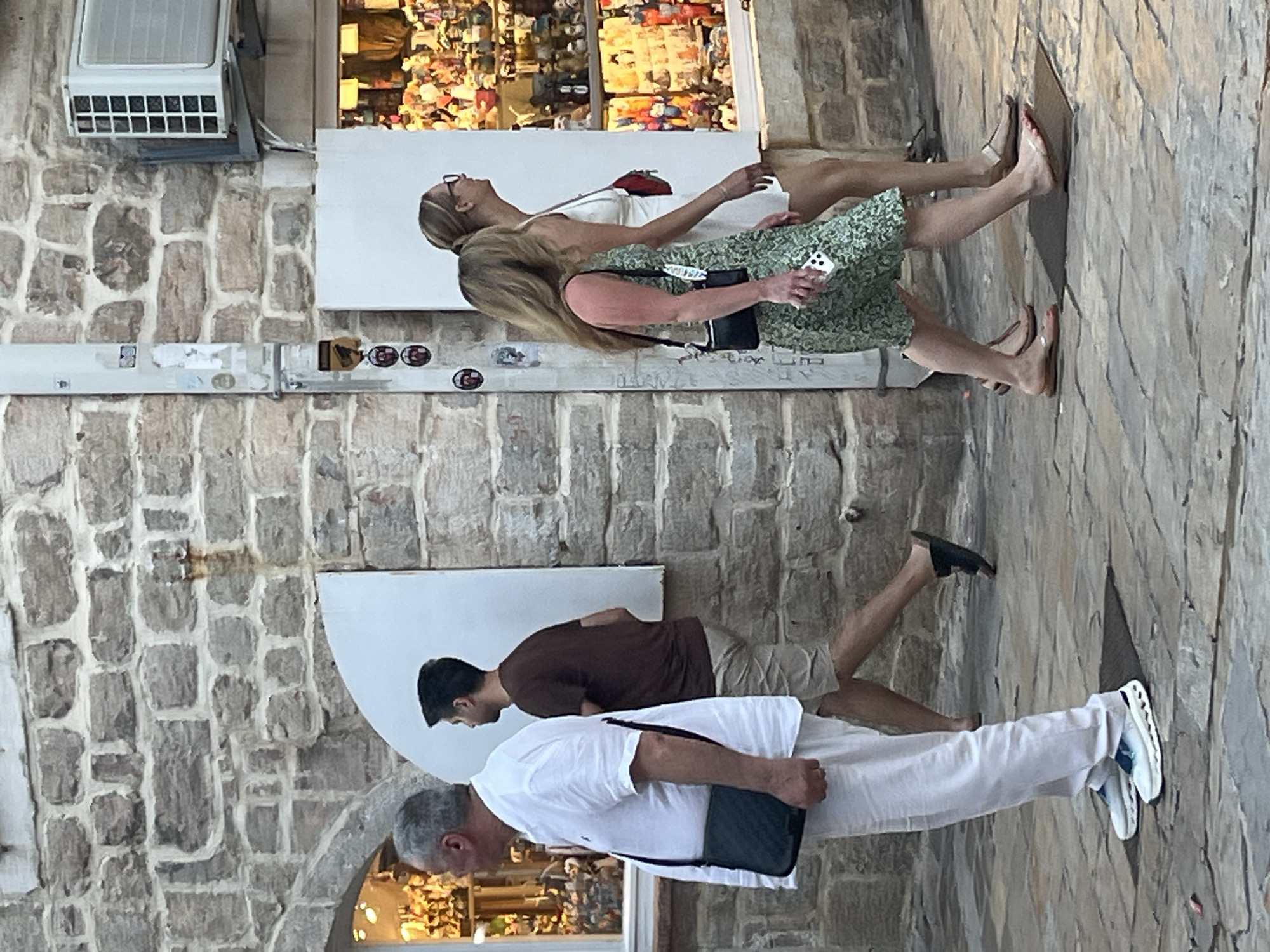 The image depicts a street scene in Budva, Montenegro. In the foreground, a man wearing a white collared shirt and white pants walks from left to right, carrying a black messenger bag across his chest. To his right, a man in a brown t-shirt and beige shorts walks away from the camera. Further to the right, two women are walking. The woman closer to the camera wears a green floral print dress and carries a black handbag. She is holding a smartphone in her right hand. The woman behind her wears a white dress and carries a red bag. They are walking past a shop window filled with merchandise. The buildings in the background are constructed with stone blocks, and an air conditioning unit is visible mounted on the wall of one of the buildings. The ground is paved with stone tiles.