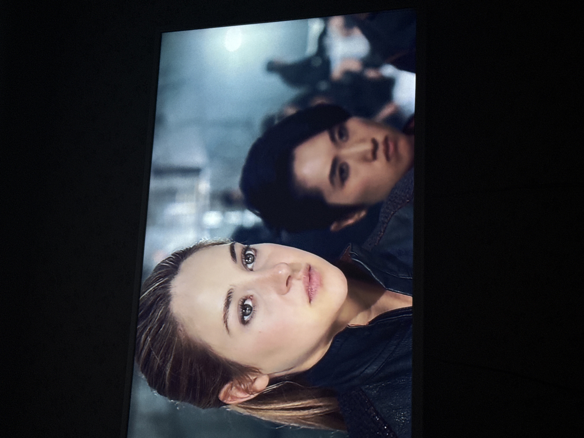The image shows a scene displayed on a television screen. Two people are in the foreground of the screen's image. A fair-skinned woman with light brown hair and brown eyes is in the front. Behind her is a man with dark hair and brown eyes. Both are wearing dark clothing that appears to be leather or a similar material. The background is blurry and shows other people, as well as some indistinct structures that could be part of a building. The rest of the image is black, framing the screen. The location is unspecified in the image.
