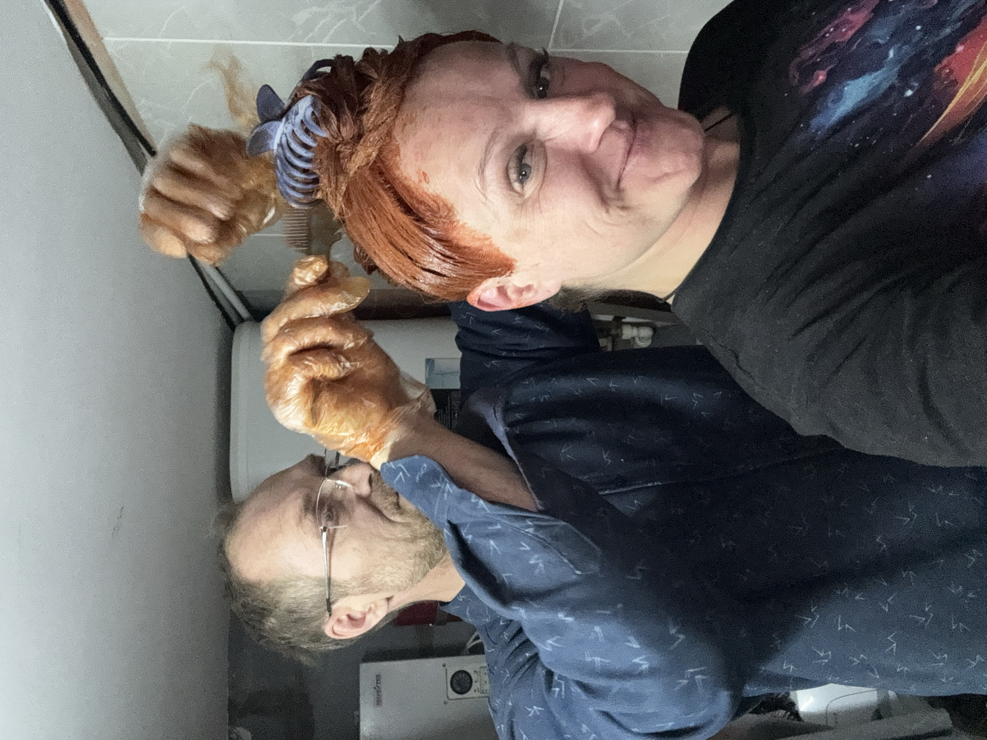 A selfie-style, indoor photograph depicts two individuals. One person, likely female, is at the top of the frame, facing the camera with a slight smile. Their hair is reddish-orange, likely from recent dye application, and partly held up with a blue hair clip. They are wearing a dark-colored top with a galaxy print. The other person, likely male, is located in the bottom half of the frame, wearing glasses and a blue shirt with a small white pattern. They are holding a brush or applicator with a gloved hand, seemingly assisting with the hair dyeing process. Both individuals have protective gloves on. The background reveals a tile wall, pipes, and a white appliance (possibly a water heater). The location is inferred to be in a domestic setting in the city of Chaikovsky, Russia, based on the image context.